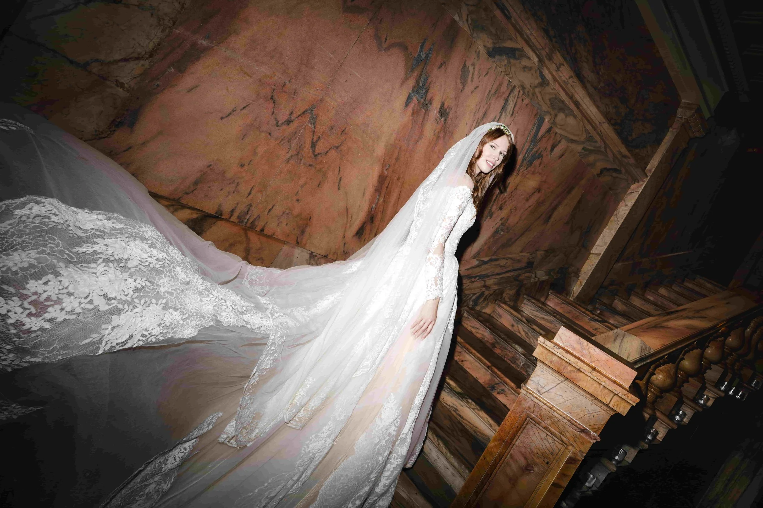 A bride in a white lace wedding gown with a long train, standing on a wooden staircase next to a marble wall, smiling.