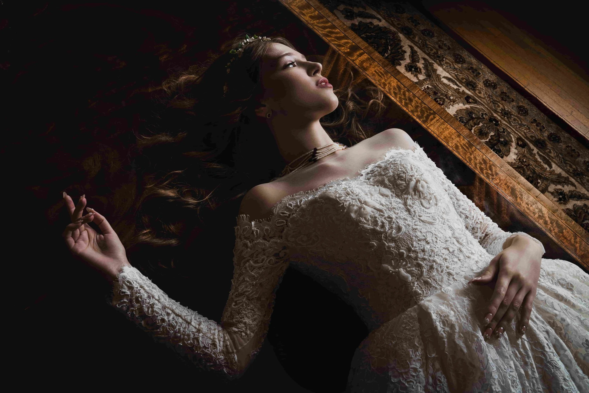 A woman in a white lace wedding dress lying on the floor with her eyes closed and an expression of calm, her hand resting on her stomach and her other hand near her head. She has long brown hair and is wearing a pearl necklace and a floral headpiece,