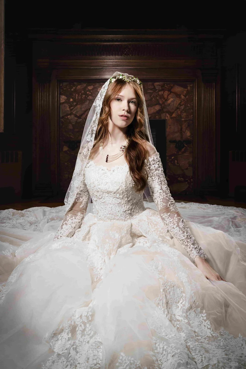 Young woman with red hair wearing a wedding dress with lace details, long sleeves, and a veil, sitting on the floor in a room with wooden paneling and a stone fireplace.