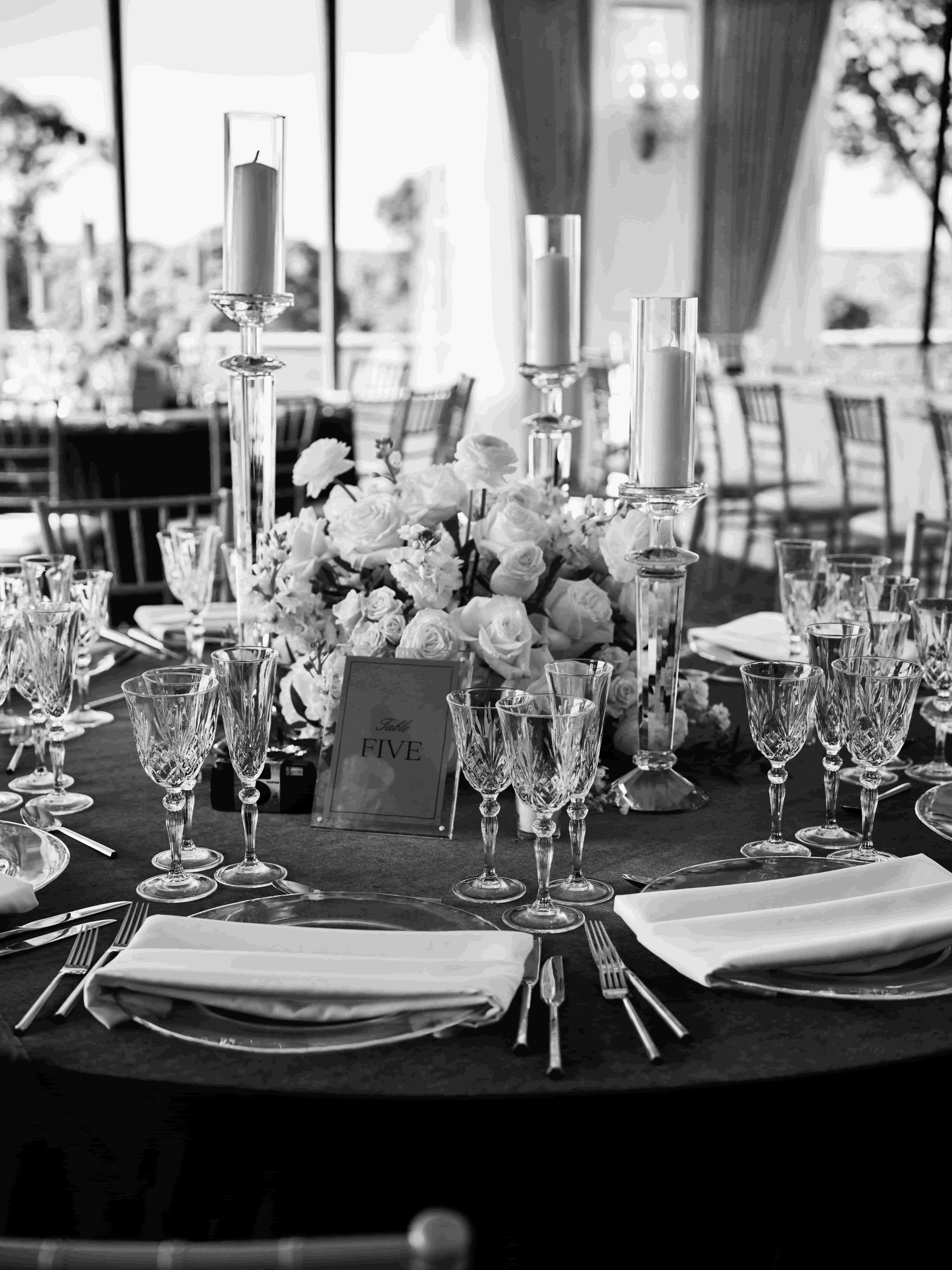 Elegant banquet table set with glassware, white napkins, floral centerpiece, and tall candles, with a table number 'Five' sign, in a bright, decorated event space.