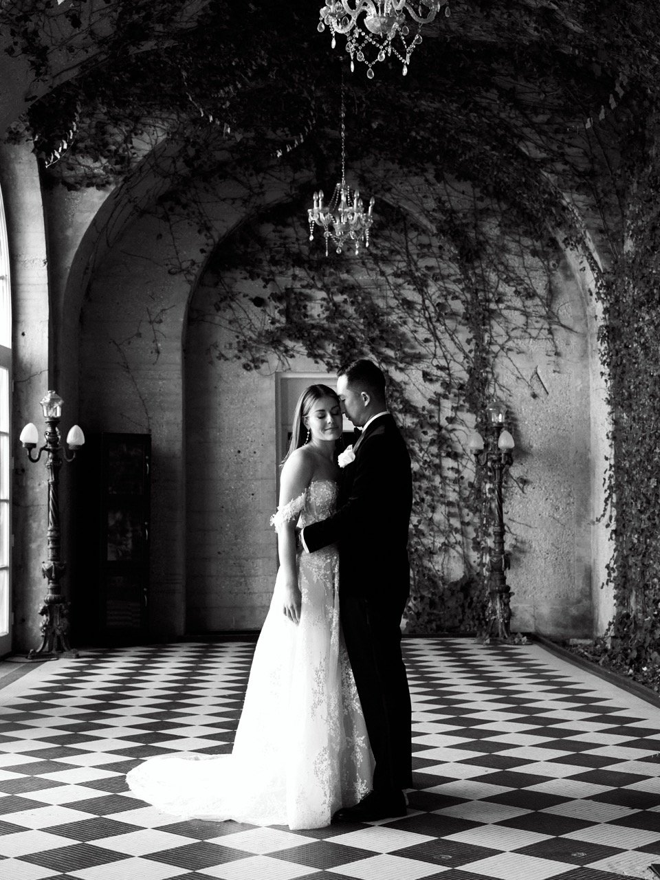 Black and white photo of a bride and groom standing close together, holding hands, in an elegant indoor setting with a checkered floor, chandeliers, and decorative lighting.