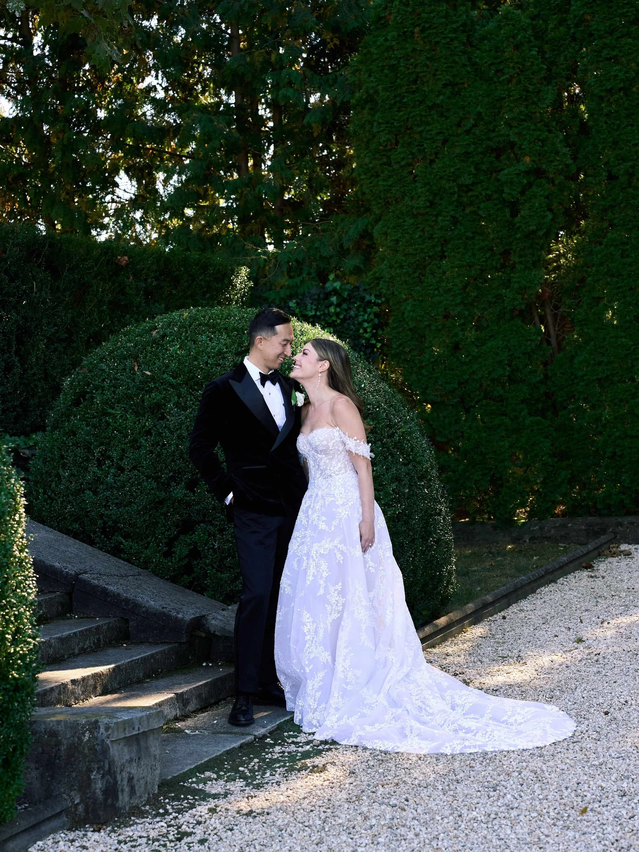 A newlywed couple stands together outdoors, dressed in wedding attire, smiling and leaning close to each other, with lush green bushes and trees in the background.
