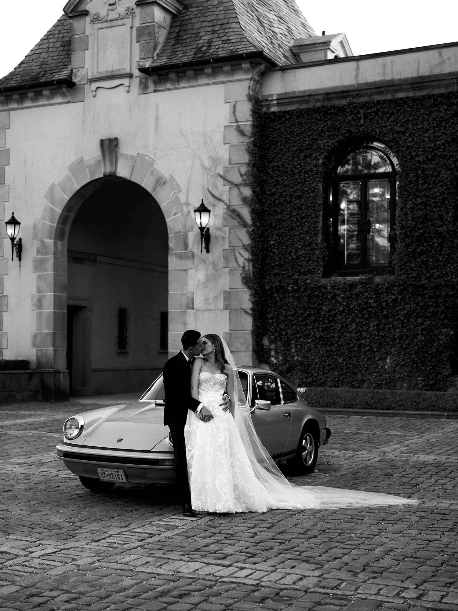 A bride and groom sharing a kiss in front of a vintage car on a cobblestone street outside a historic building with an arched entrance and ivy-covered walls, black and white photo.