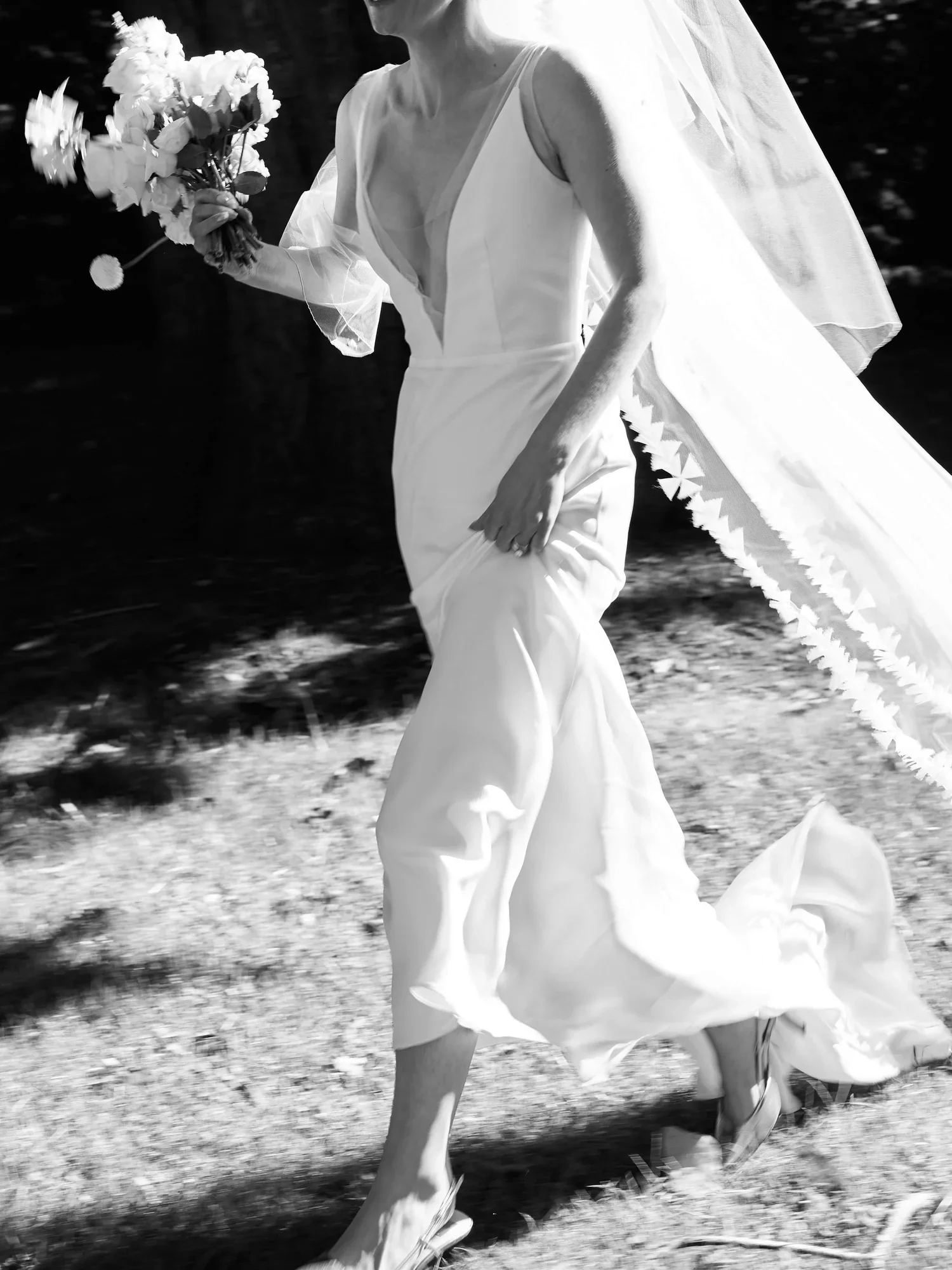 A woman in a wedding dress holding a bouquet of flowers, walking outdoors in sunlight.