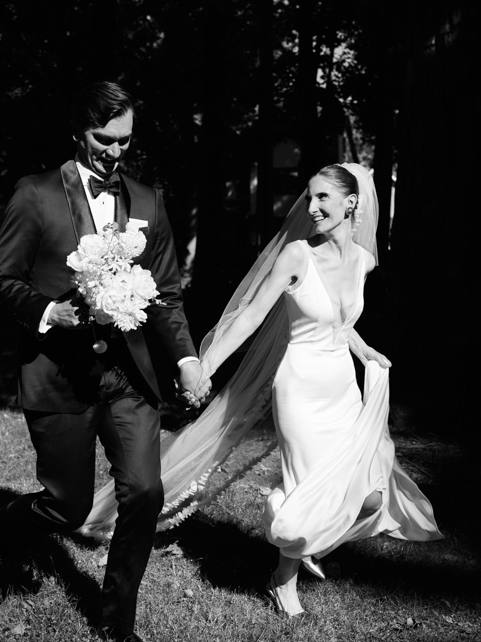 A bride and groom walking outdoors on their wedding day, holding hands, with the bride wearing a long white dress and veil, and the groom in a tuxedo holding a bouquet of flowers.