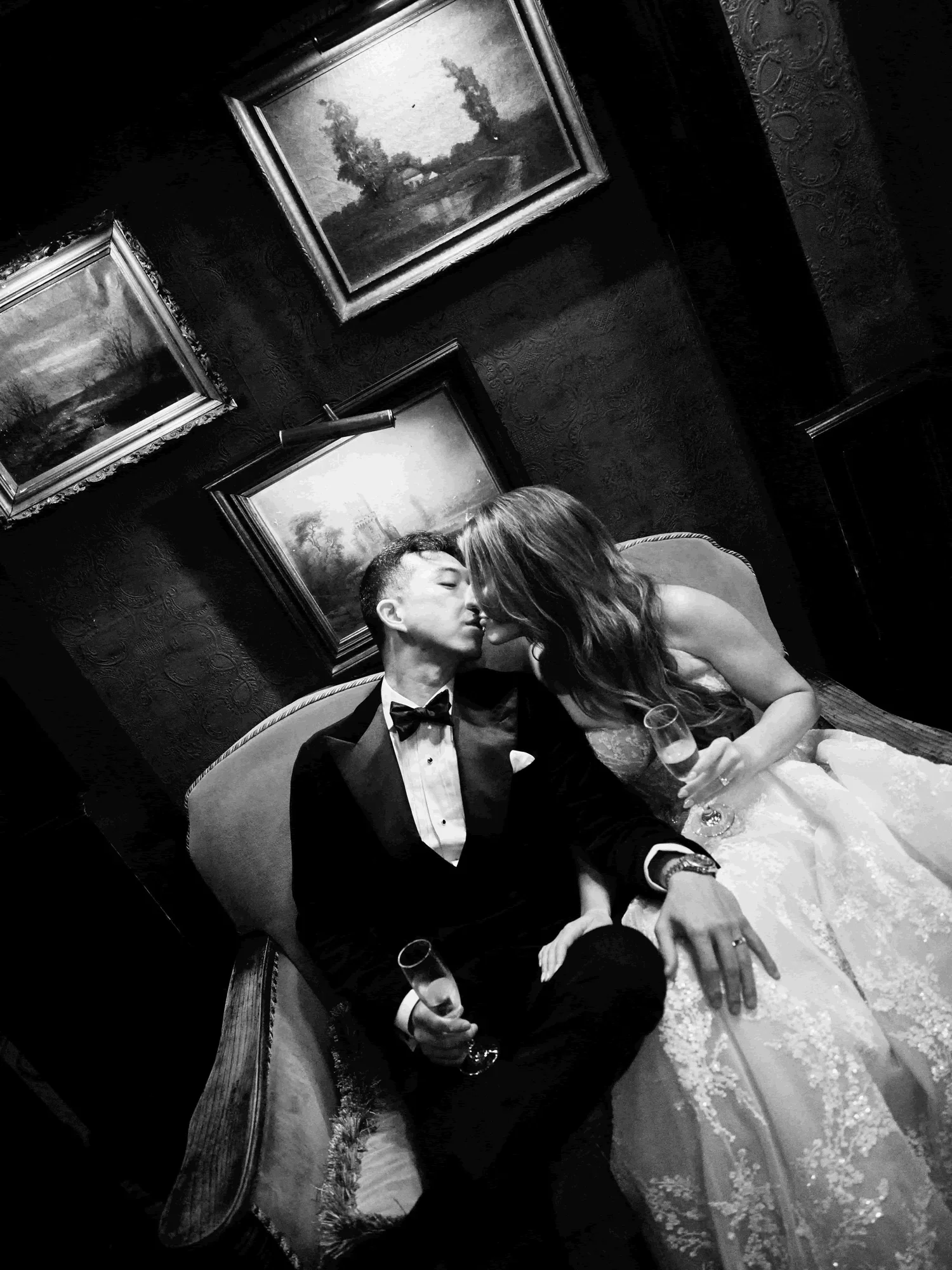A black and white photo of a couple sitting on a vintage sofa, about to kiss, holding champagne glasses. The man is in a tuxedo, and the woman is in a wedding dress. Several framed landscape paintings hang on the dark patterned wall behind them.