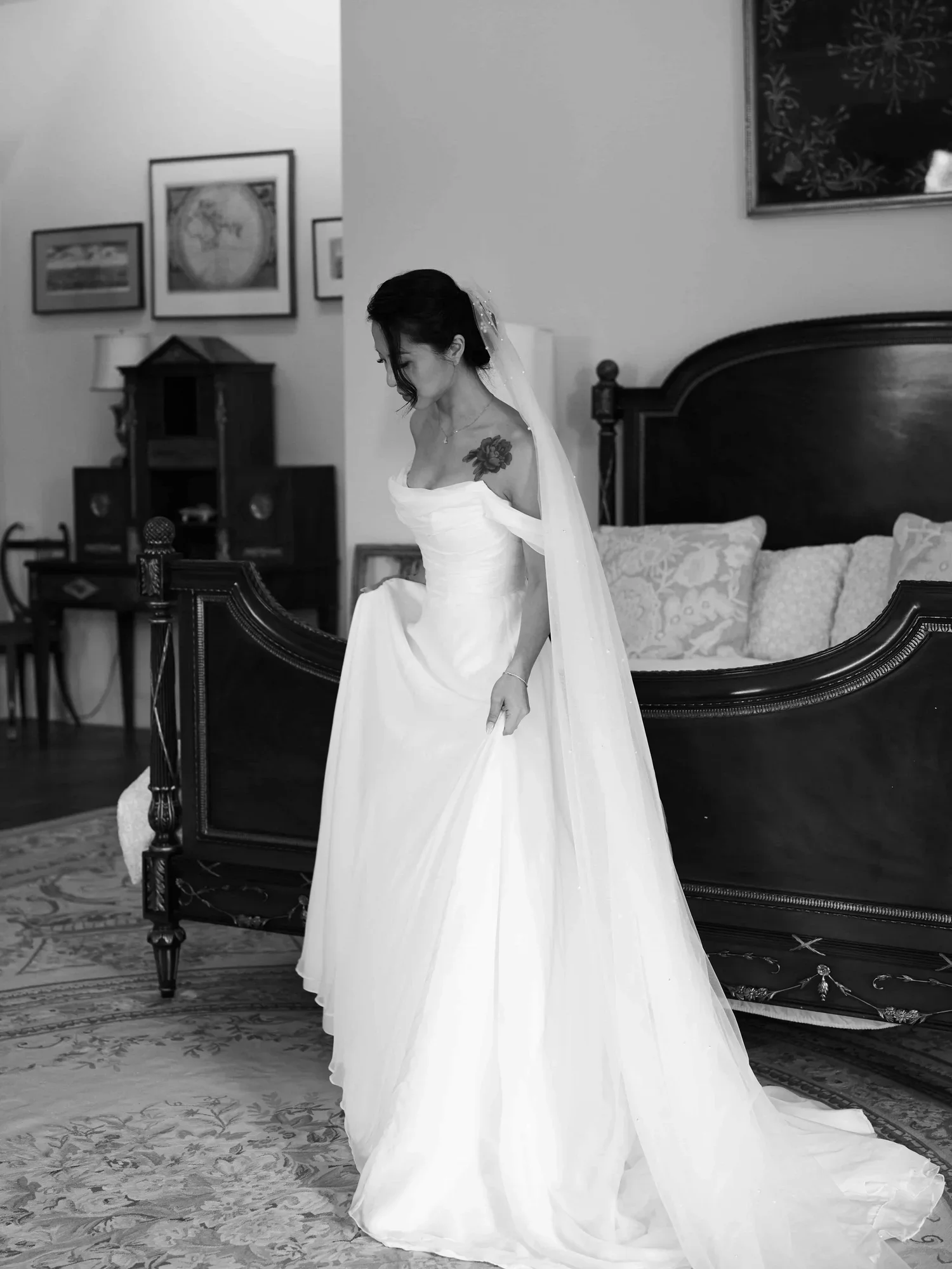 A woman in a white wedding dress and veil, standing in a living room, looking down and holding the edge of her dress.
