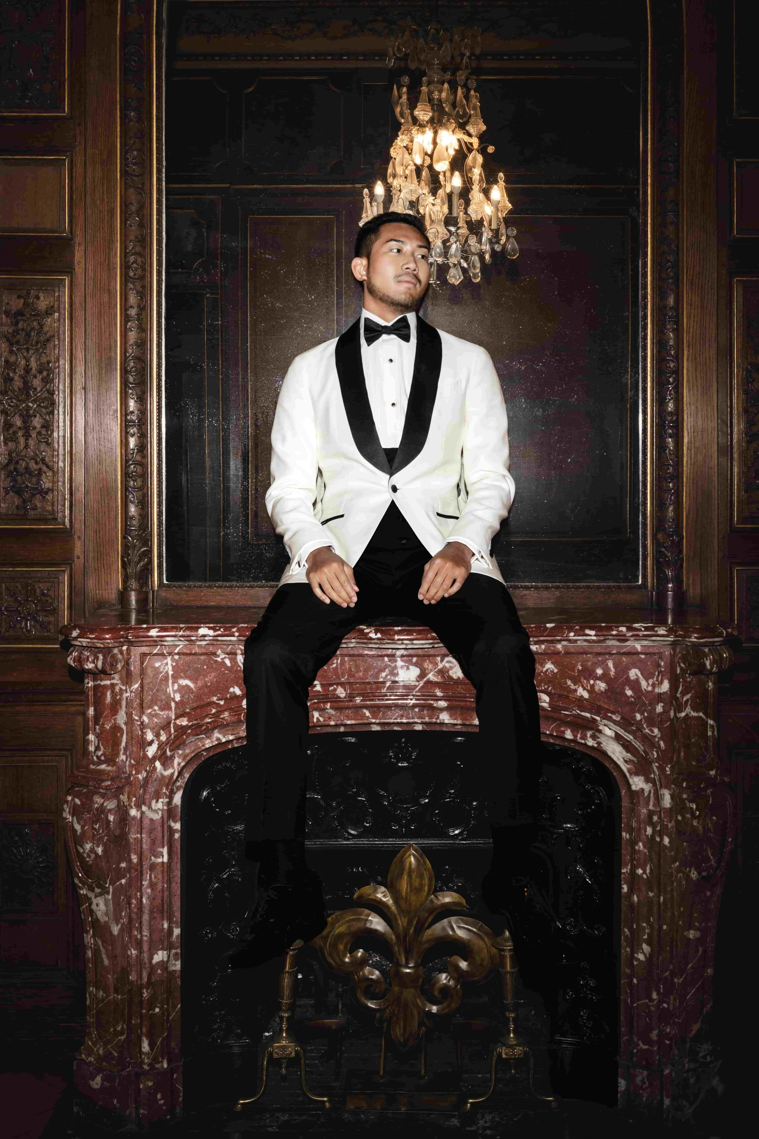 A groom wearing a tuxedo with a white jacket and black lapels seated on a marble fireplace mantel in an elegant room with dark wood paneling and a chandelier overhead.