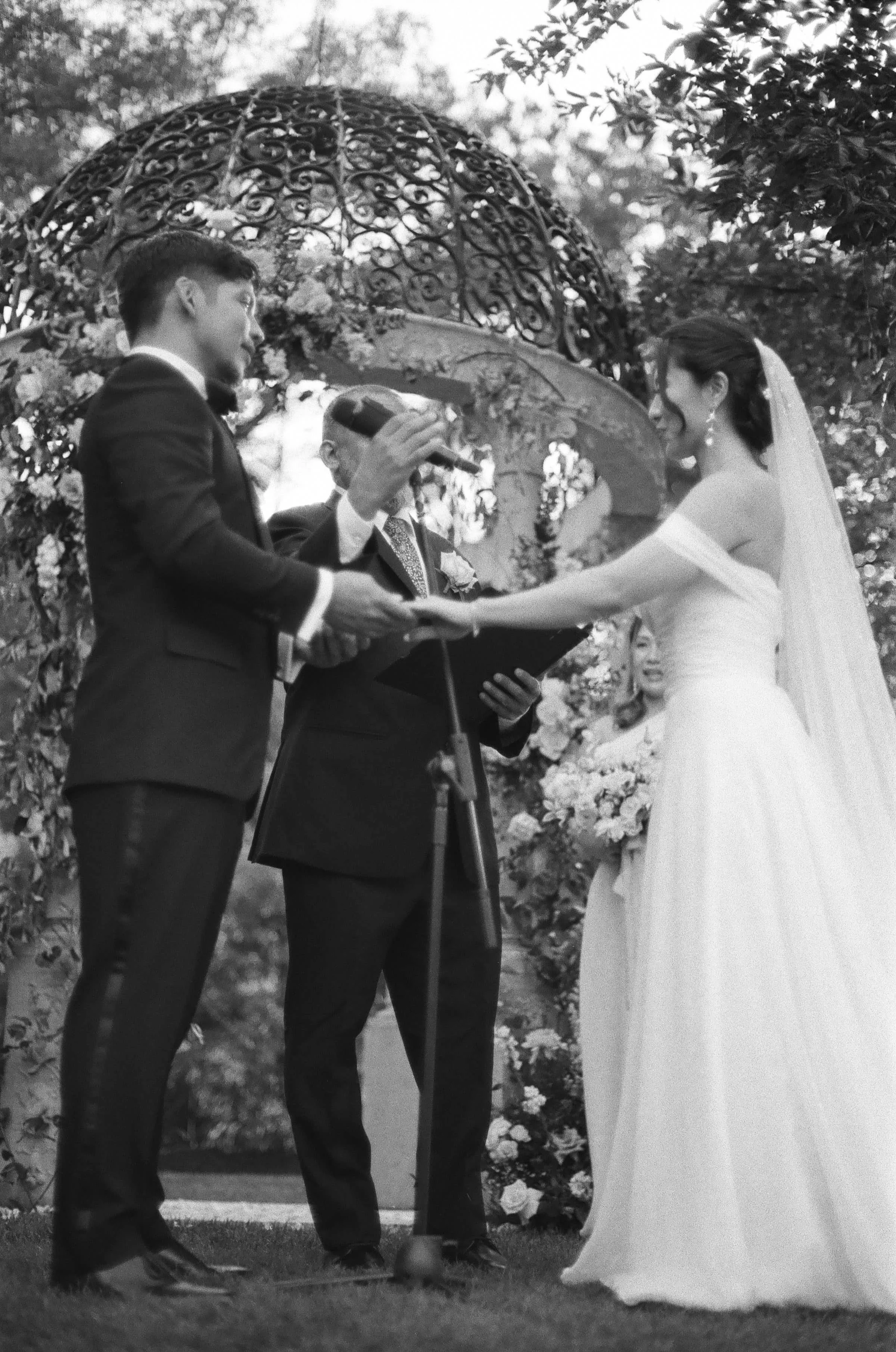 A black and white photo of a wedding ceremony with a bride and groom holding hands, an officiant reading, and an ornate floral arch in the background.