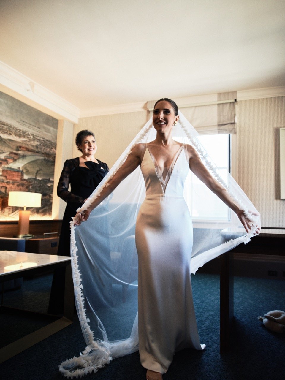 Bride in a white wedding gown with a sheer neckline, standing in a room with sunlight coming through a window, smiling, with a woman in black standing behind her.