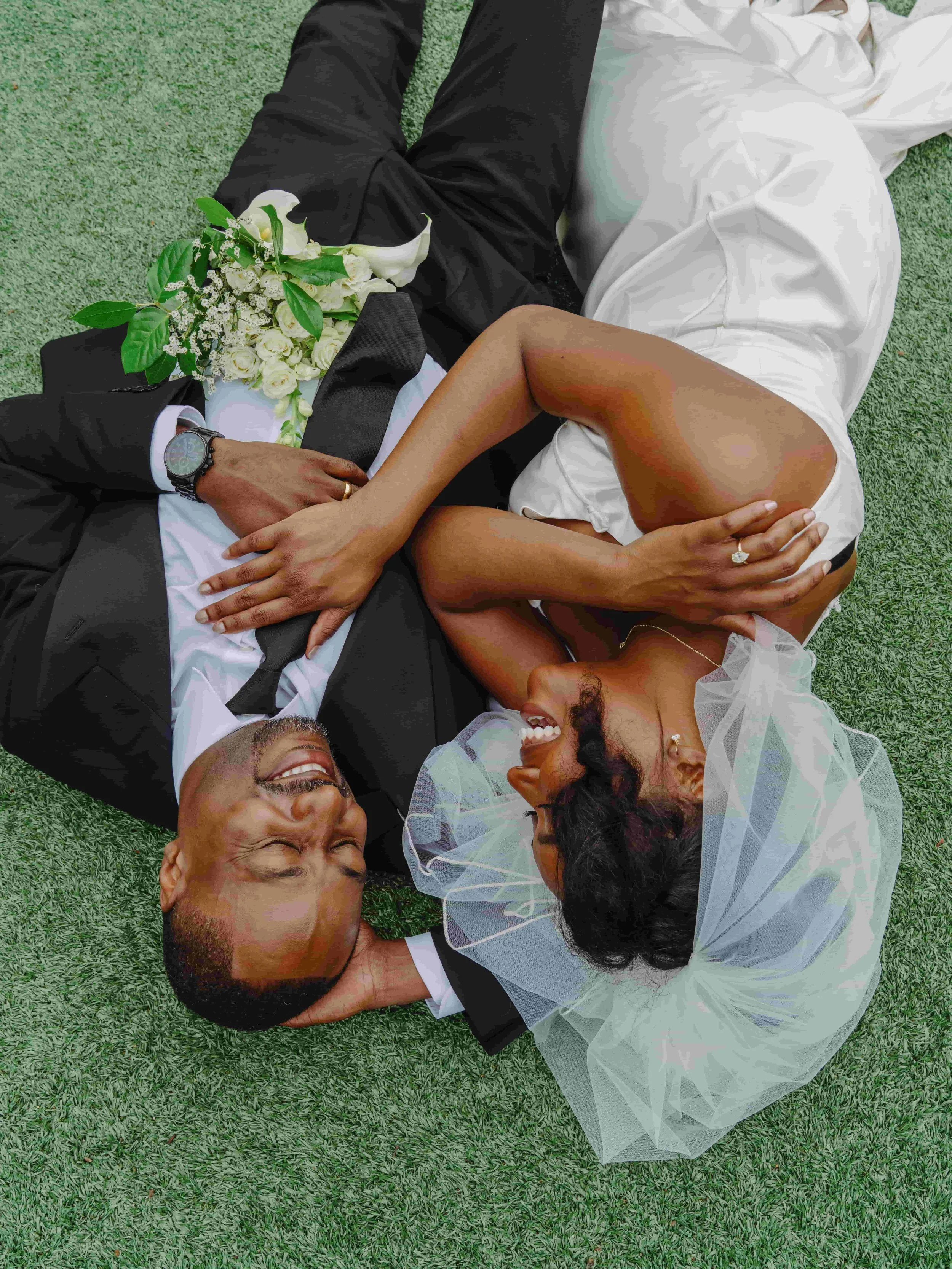 A newlywed couple lies on grass, smiling and looking at each other. The groom wears a black tuxedo with a watch and a boutonniere. The bride wears a white wedding dress, veil, and ring, holding a bouquet of white flowers.