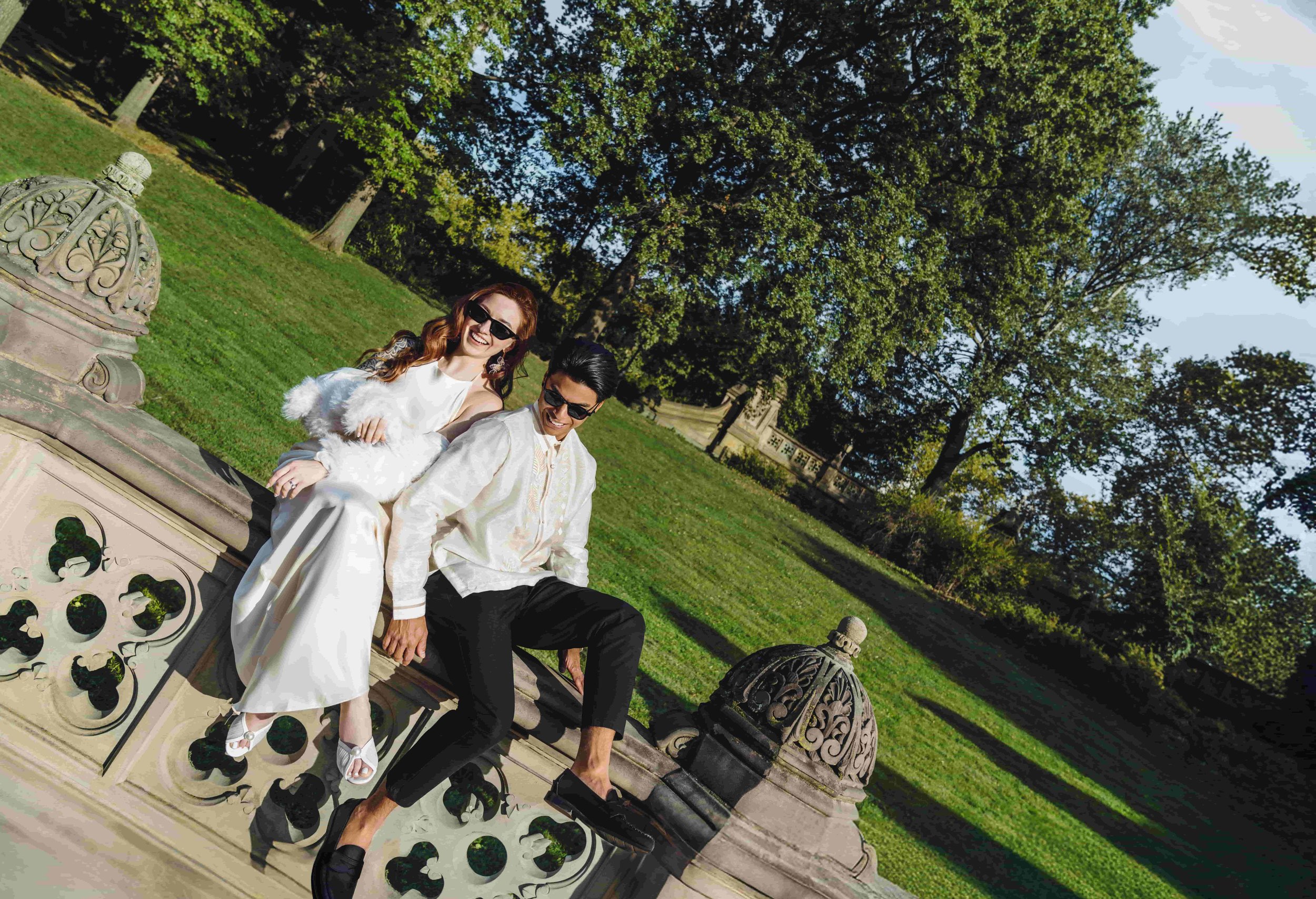 A couple sitting on a stone railing in a park with lush green trees in the background. The woman is wearing a white dress with fluffy sleeves, sunglasses, and white shoes, while the man is dressed in a cream-colored jacket, black pants, and black loa
