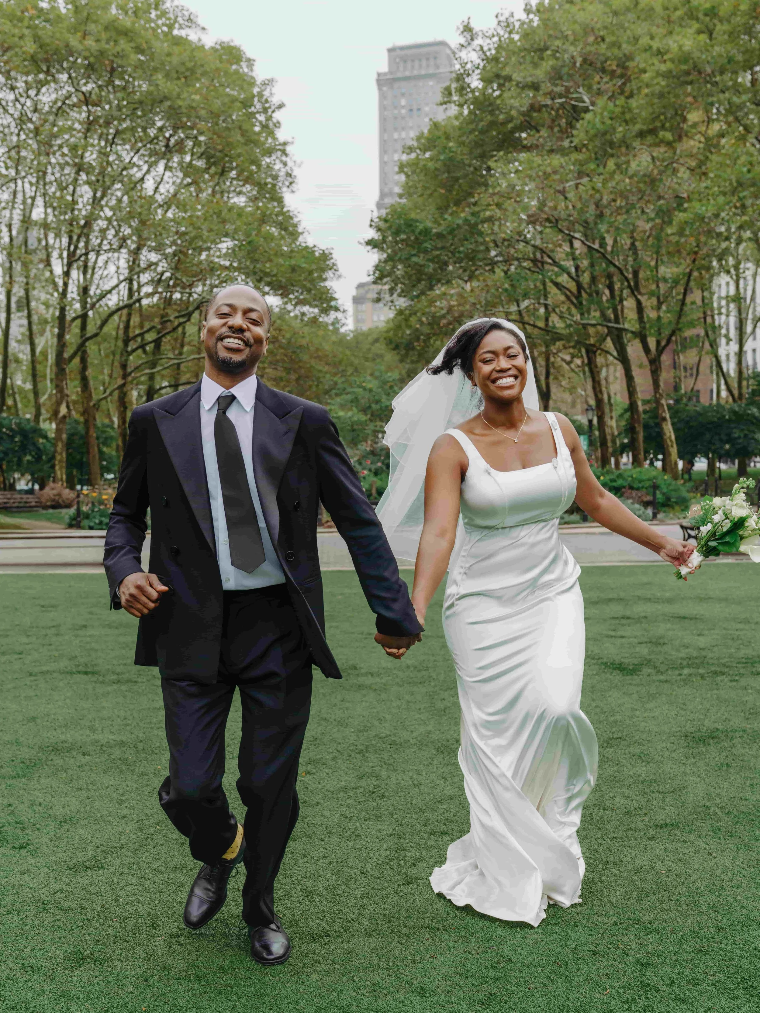 A joyful couple walking hand in hand outdoors in a park, with the woman in a white wedding dress and veil, holding a bouquet, and the man in a dark suit and tie, surrounded by trees and city buildings in the background.
