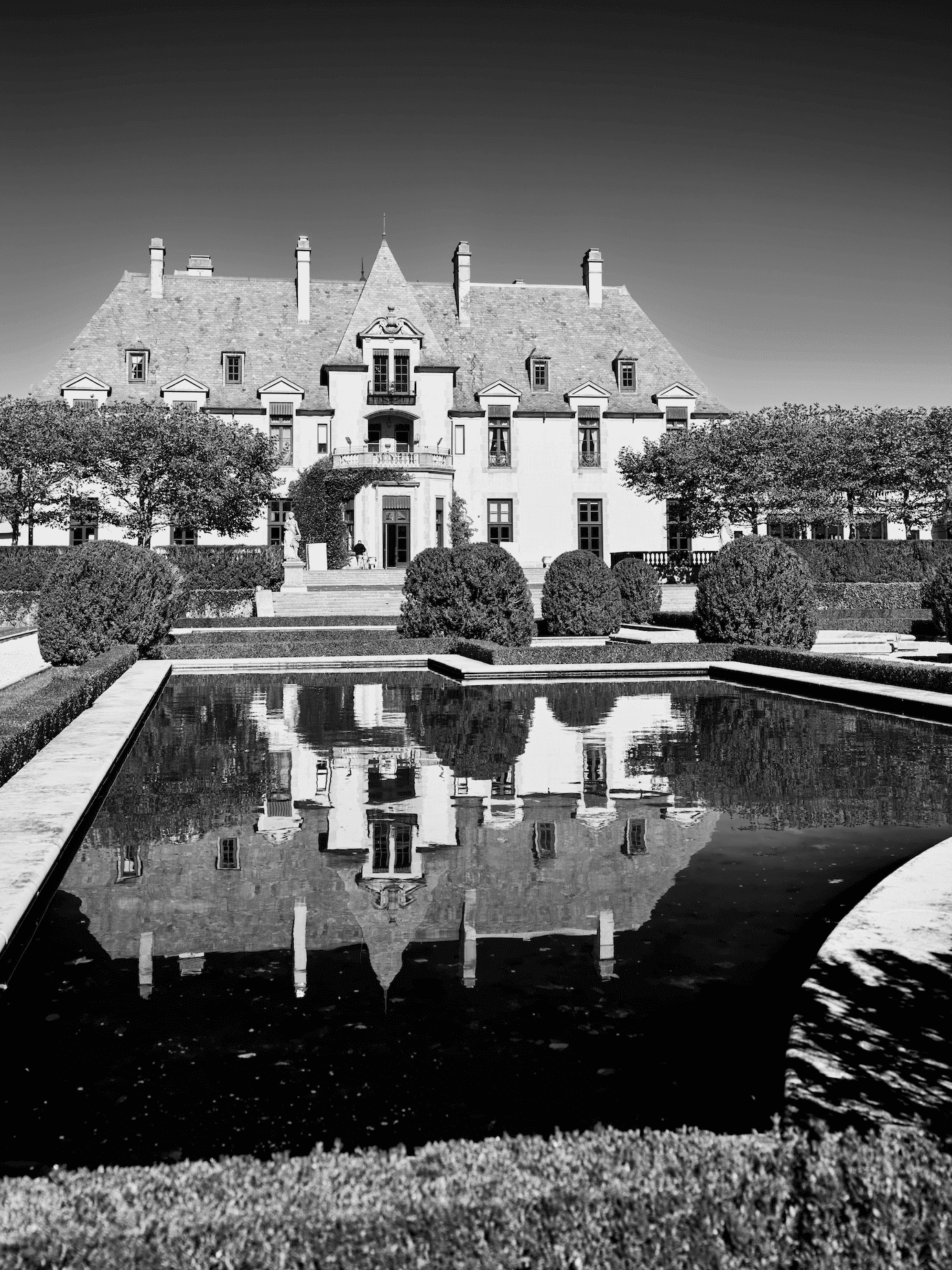 A large historic mansion with multiple stories and a steep roof, reflected in a rectangular pond in the garden, surrounded by neatly trimmed bushes and trees.