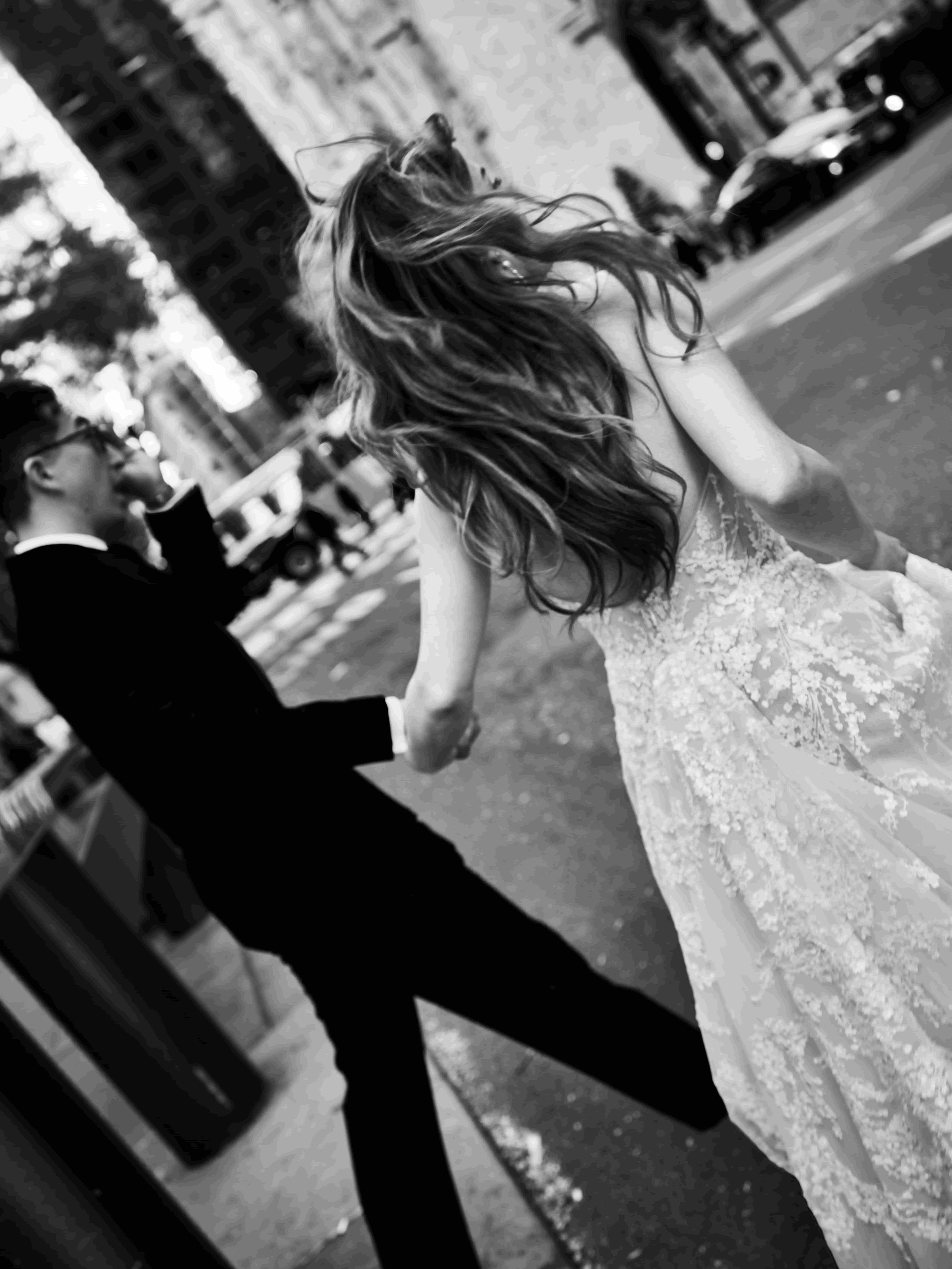 A black and white photo of a bride and groom holding hands, walking on a city street. The bride has long, wavy hair and is wearing a lace wedding dress. The groom is dressed in a black tuxedo. People and cars are visible in the background.