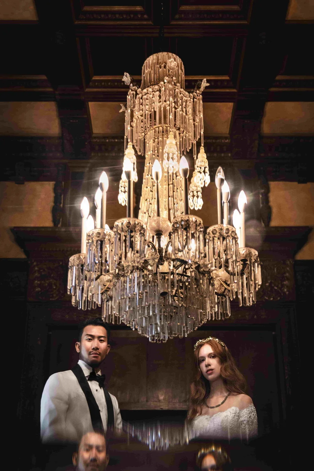 A large crystal chandelier hanging from a dark, ornate ceiling, with a man in a tuxedo and a woman in a white lace dress standing below it.
