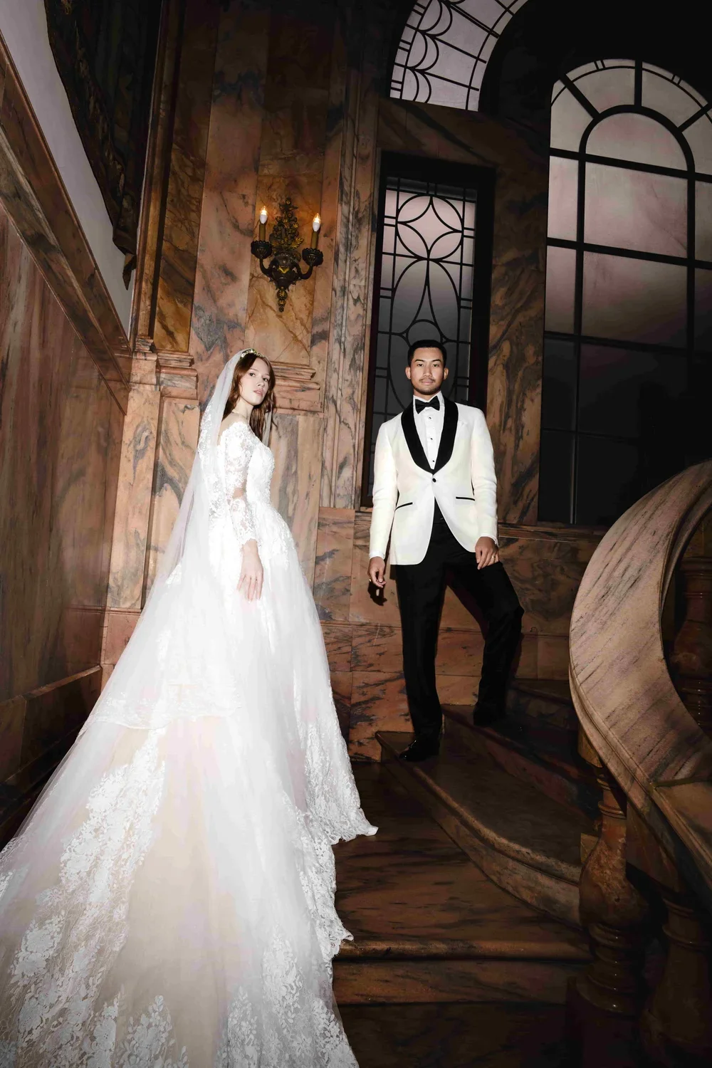 A bride in a white wedding gown and veil standing on a marble staircase next to a groom in a white tuxedo jacket with black lapels, black pants, and a black bow tie, inside an elegant, historic building with marble walls and large windows.
