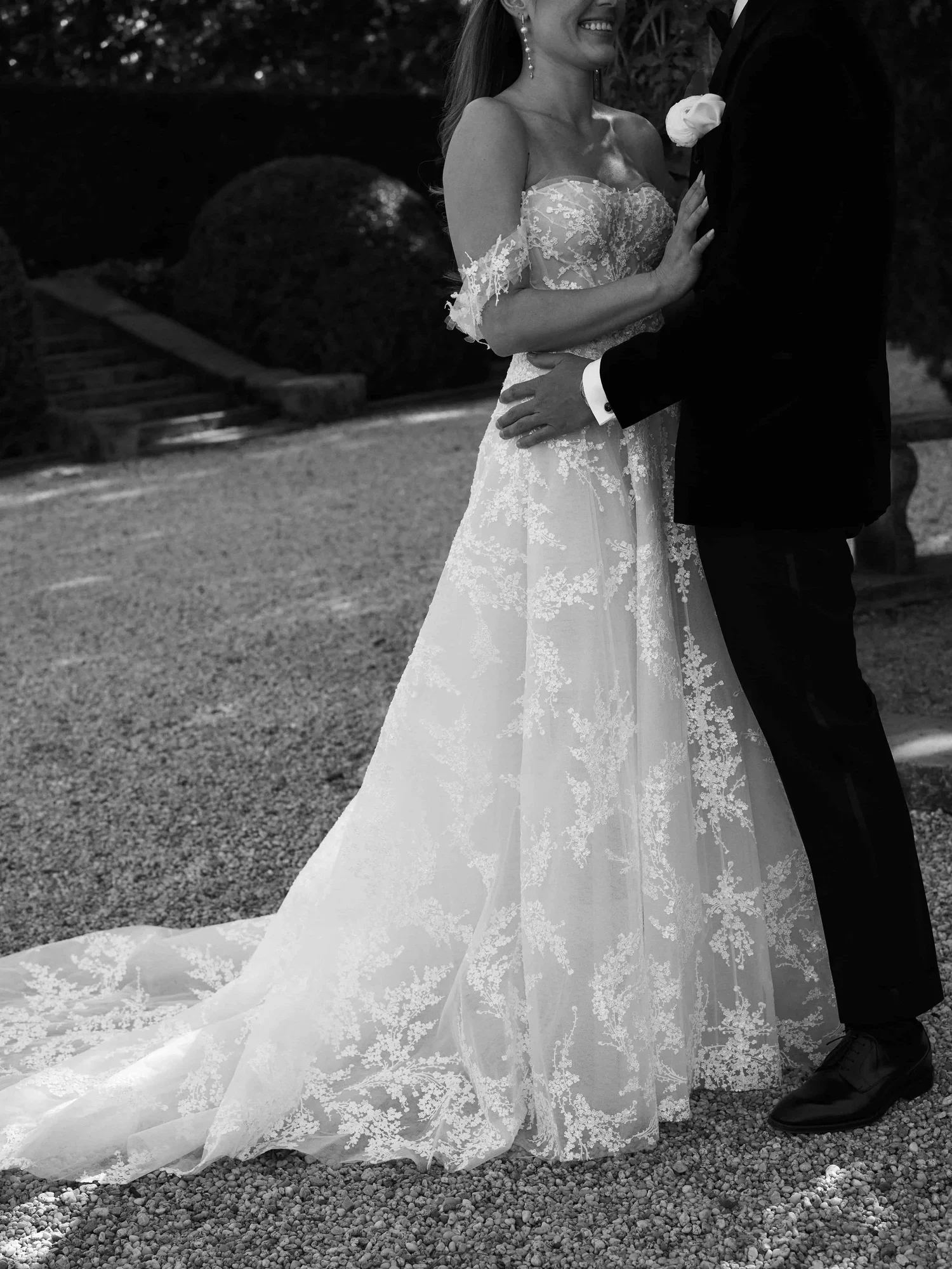 A black and white photo of a bride and groom holding each other during their wedding. The bride is wearing a lace wedding gown with off-the-shoulder sleeves, and the groom is in a black suit with a white shirt and boutonniere. They are standing outdo