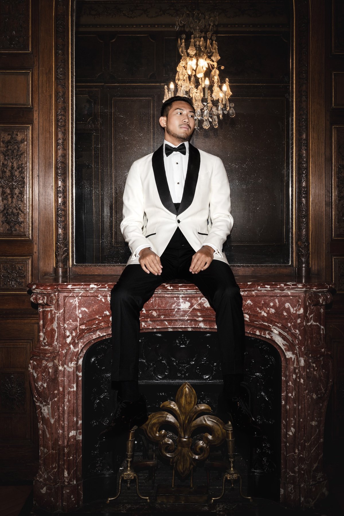 groom in a white tuxedo with black lapels and a bow tie sitting on a red marble fireplace with a chandelier hanging above him in a wood-paneled room.