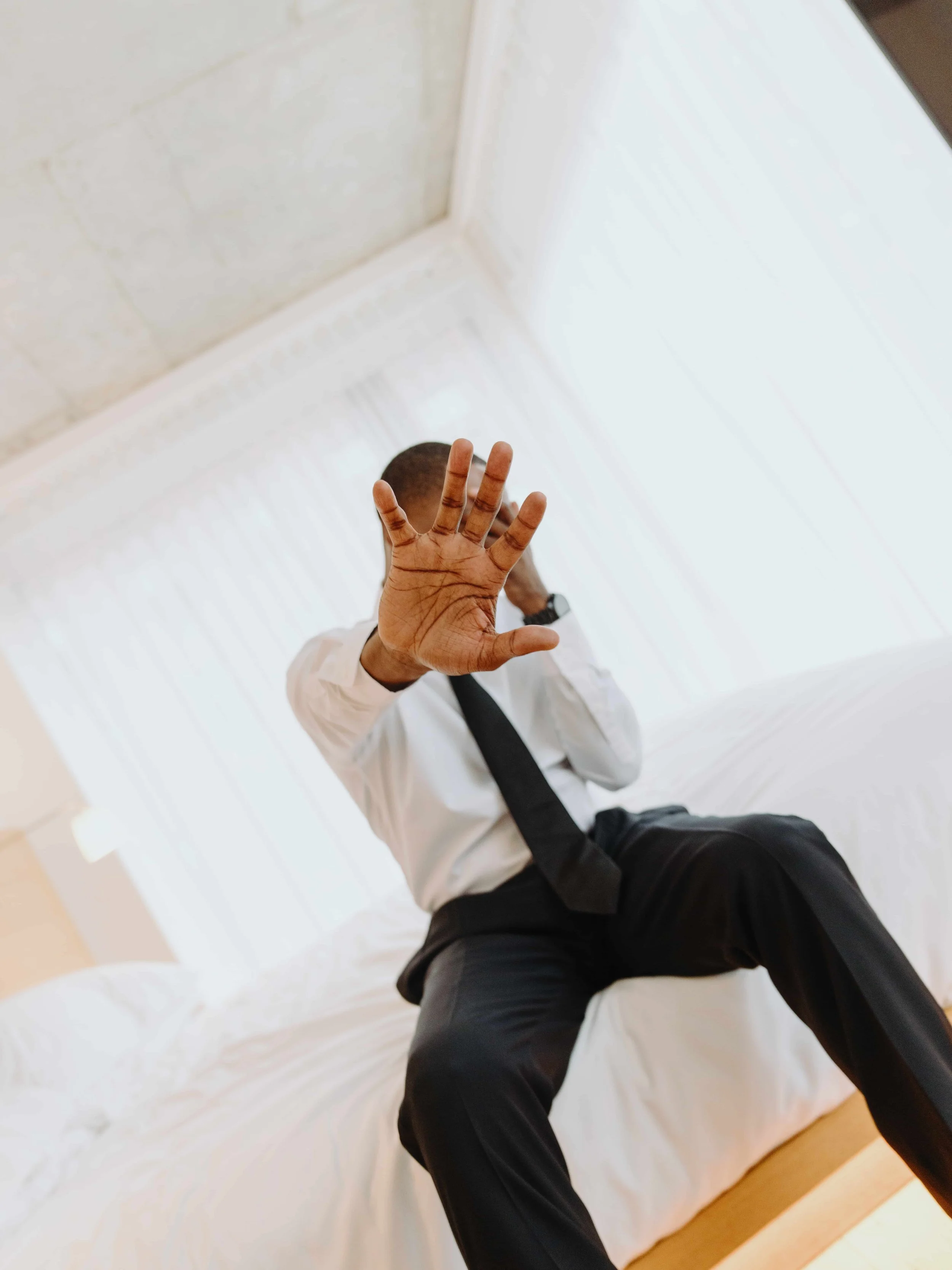 Man in business attire sitting on bed, reaching out with his hand toward the camera, obscuring his face.