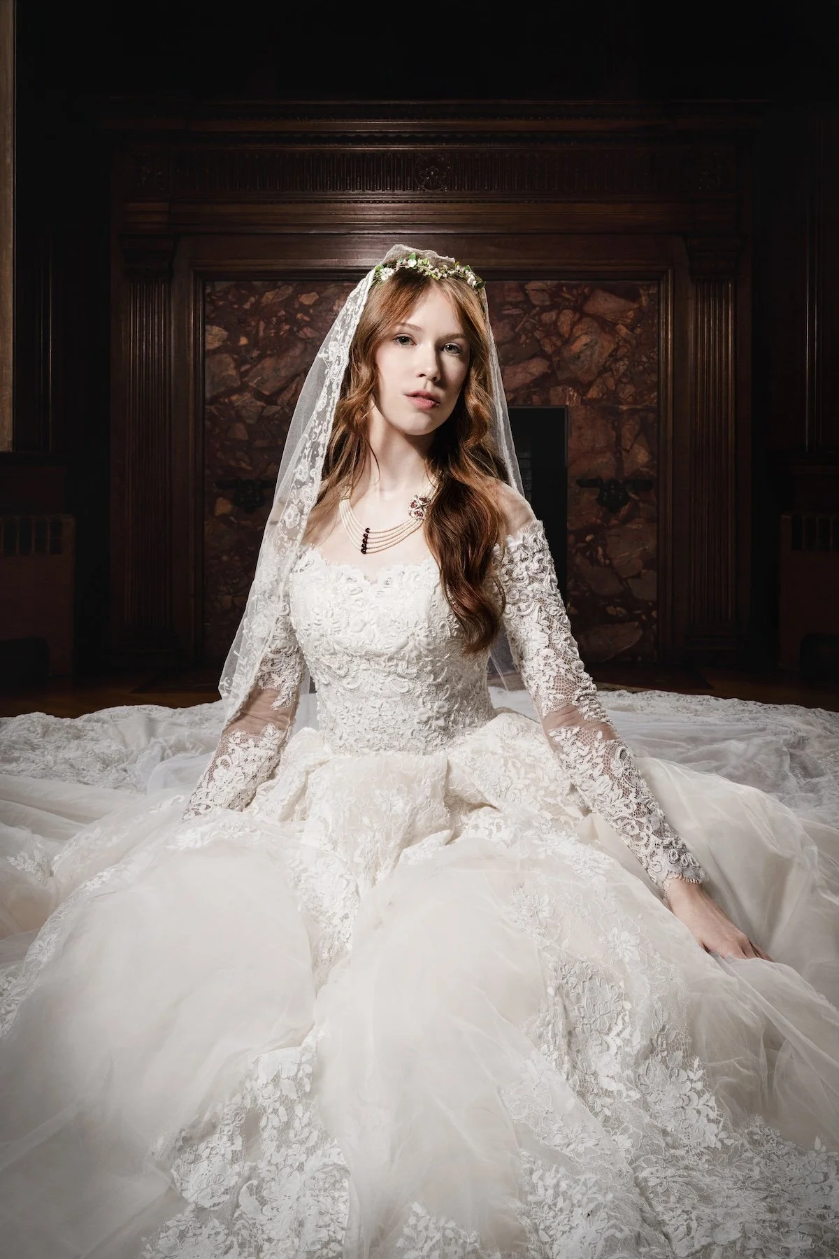 A bride in a wedding dress with lace details, sitting on the floor in an elegant room with dark wood paneling and marble wall behind her.