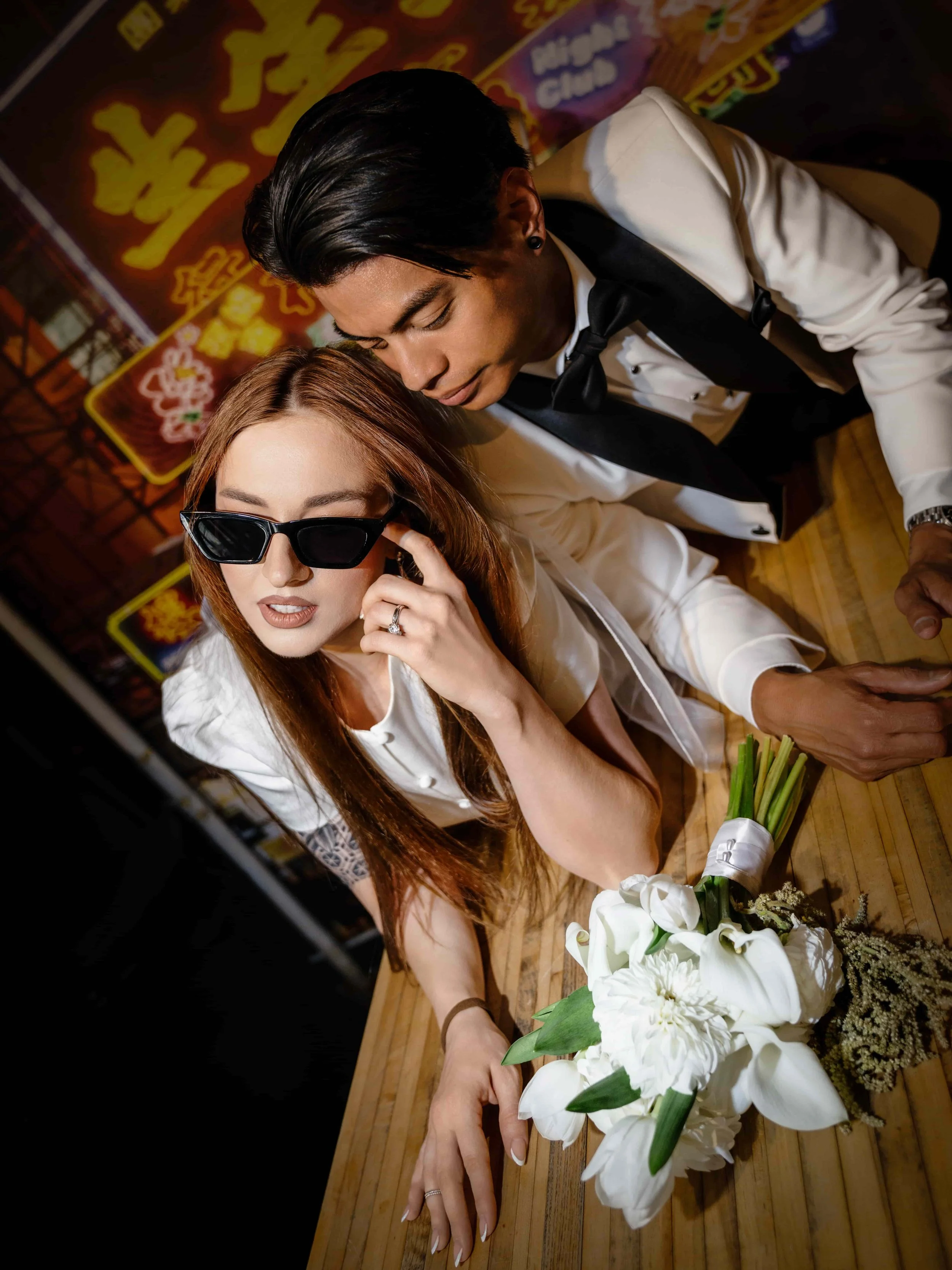 A bride in sunglasses and a white dress lying on a wooden table with a bouquet of white flowers, while a man in a tuxedo leans over her in a dimly lit setting with colorful neon signs in the background.