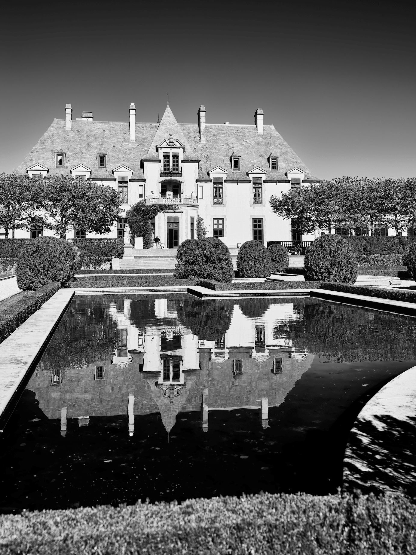 A black and white photo of a large historic mansion with a reflecting pool in front, surrounded by manicured bushes and trees.