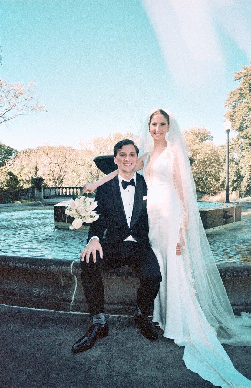 A newlywed couple posing in front of a fountain outdoors on a sunny day, with the bride in a white wedding gown and veil, and the groom in a black tuxedo, holding a bouquet of flowers.