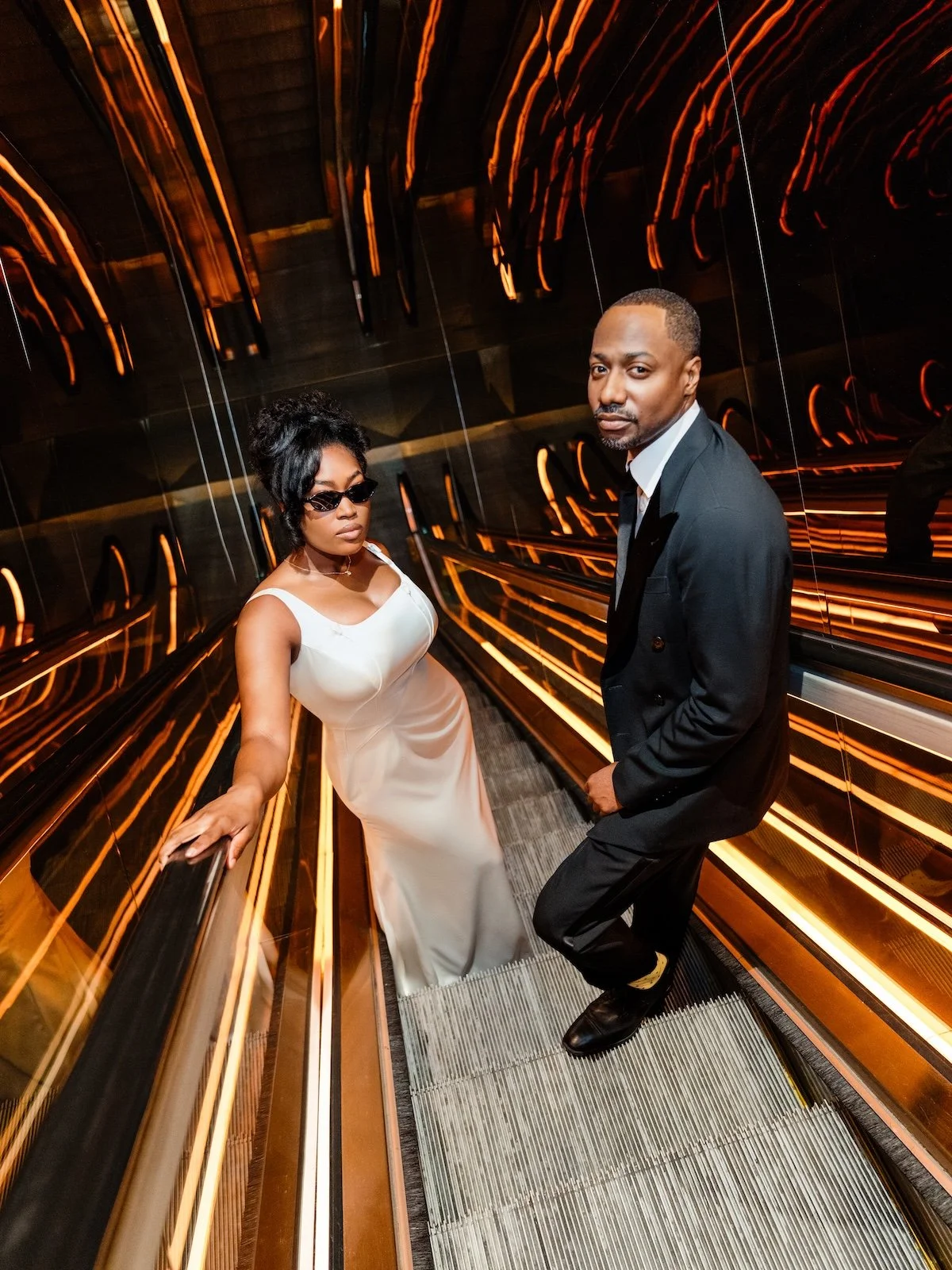 A stylish man in a black suit and white shirt stands on an escalator next to a woman in a white dress with sunglasses inside an elevator with reflective orange lighting.
