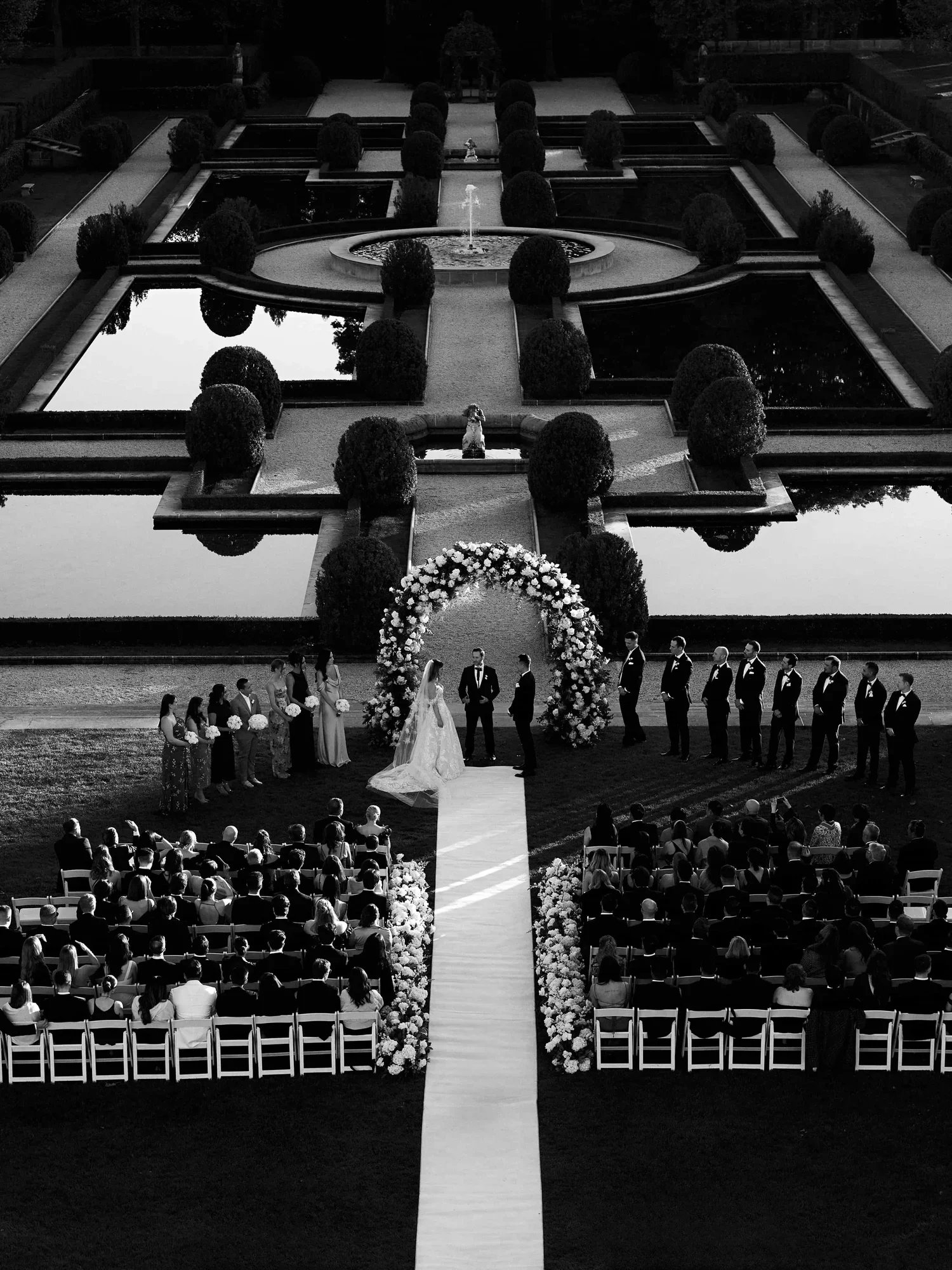 A wedding ceremony taking place outdoors with a bride and groom, officiant, bridal party, and guests seated in chairs, with a floral arch and a large formal garden with reflecting pools and a fountain in the background.