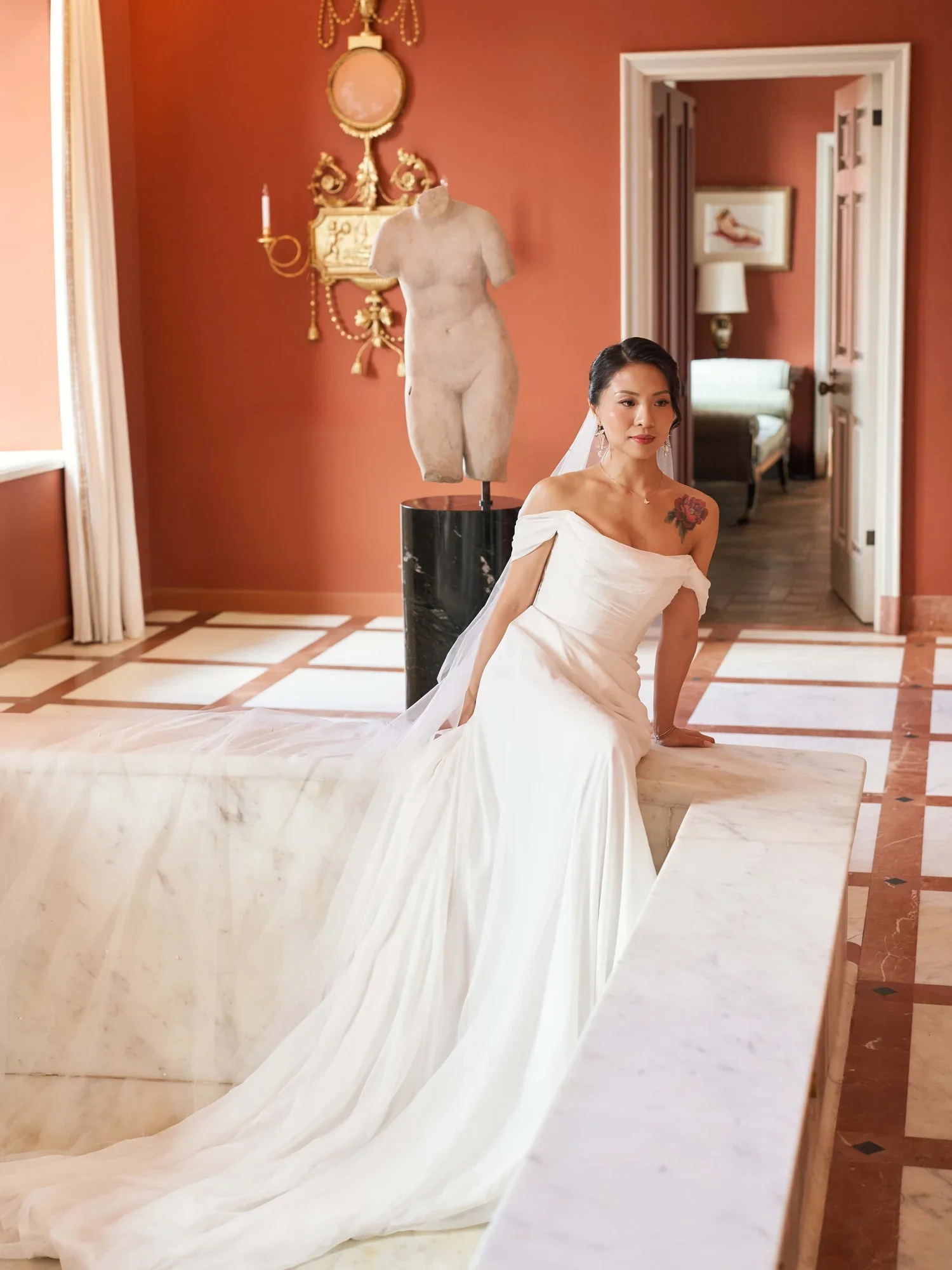 A woman in a white wedding dress sitting on a marble ledge in a richly decorated room with pink walls, a classical statue, and a doorway leading to another room with furniture and a framed picture.