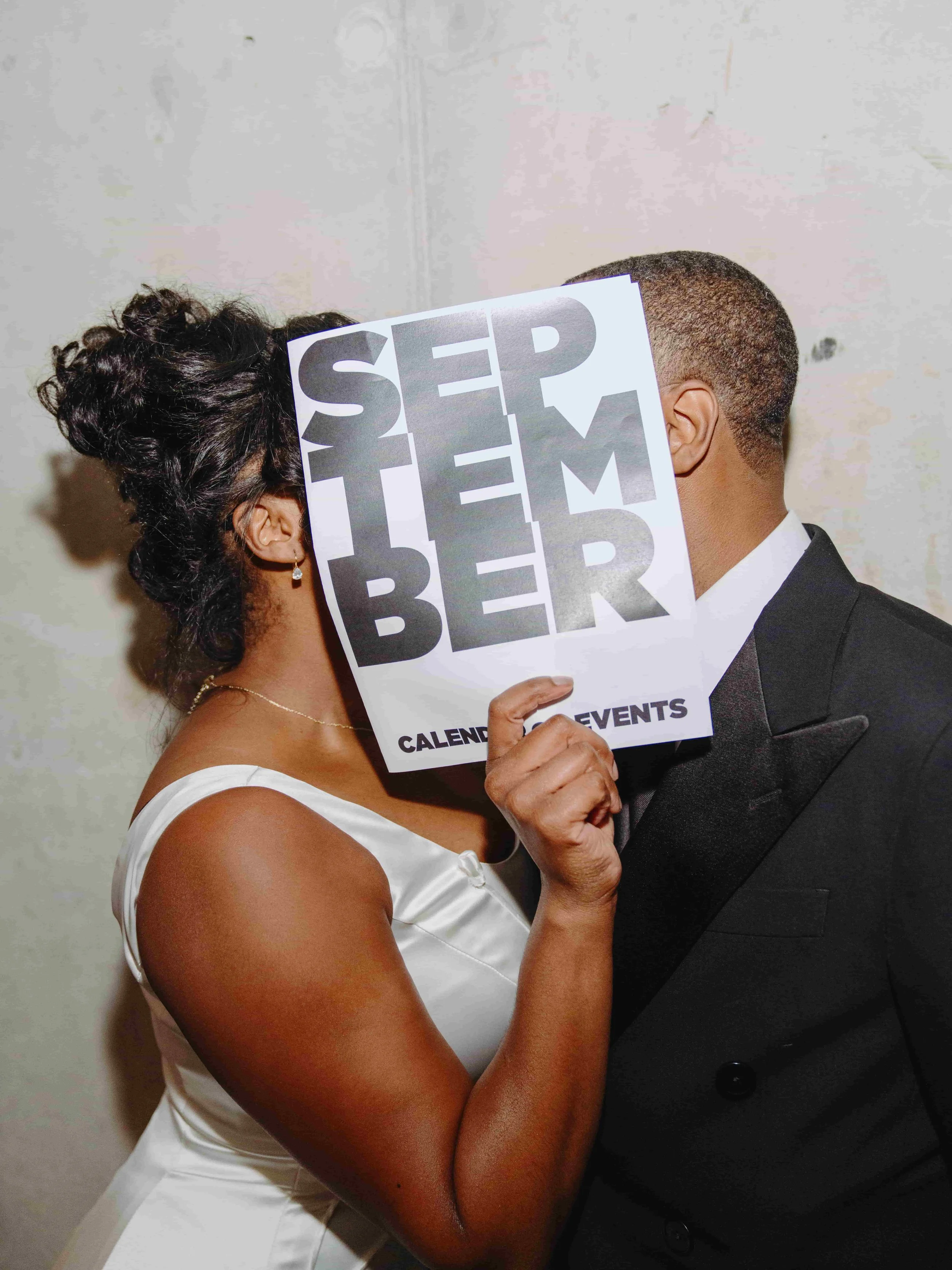 A  in a white dress and a man in a black tuxedo face each other, with the woman's arm around the man's neck. The woman's hair is styled in curls, and she is wearing earrings and a necklace. The woman is holding a sign in front of her face that says '