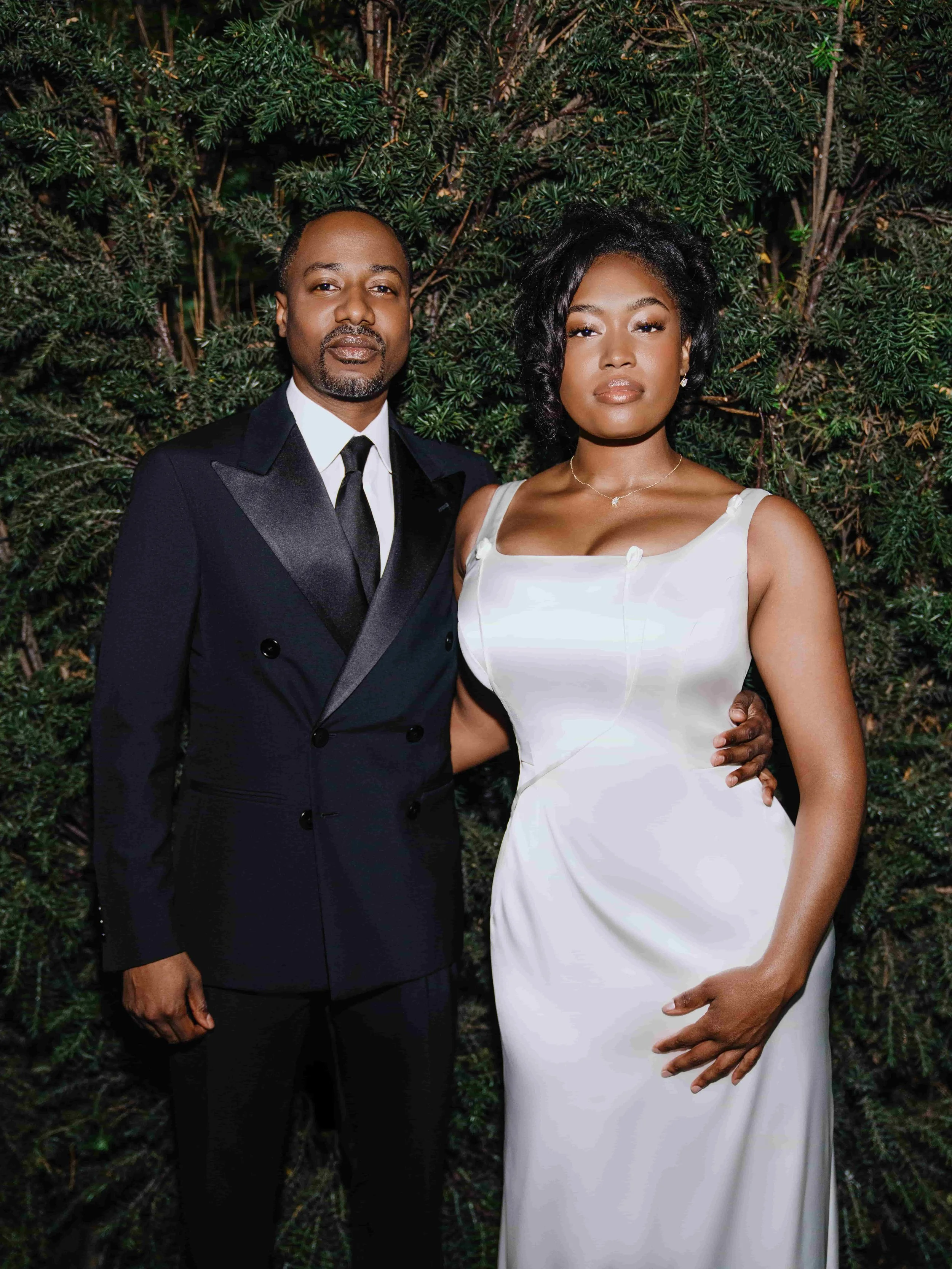 A groom in a dark tuxedo and a bride in a white gown standing together in front of green foliage, posing for a photograph.