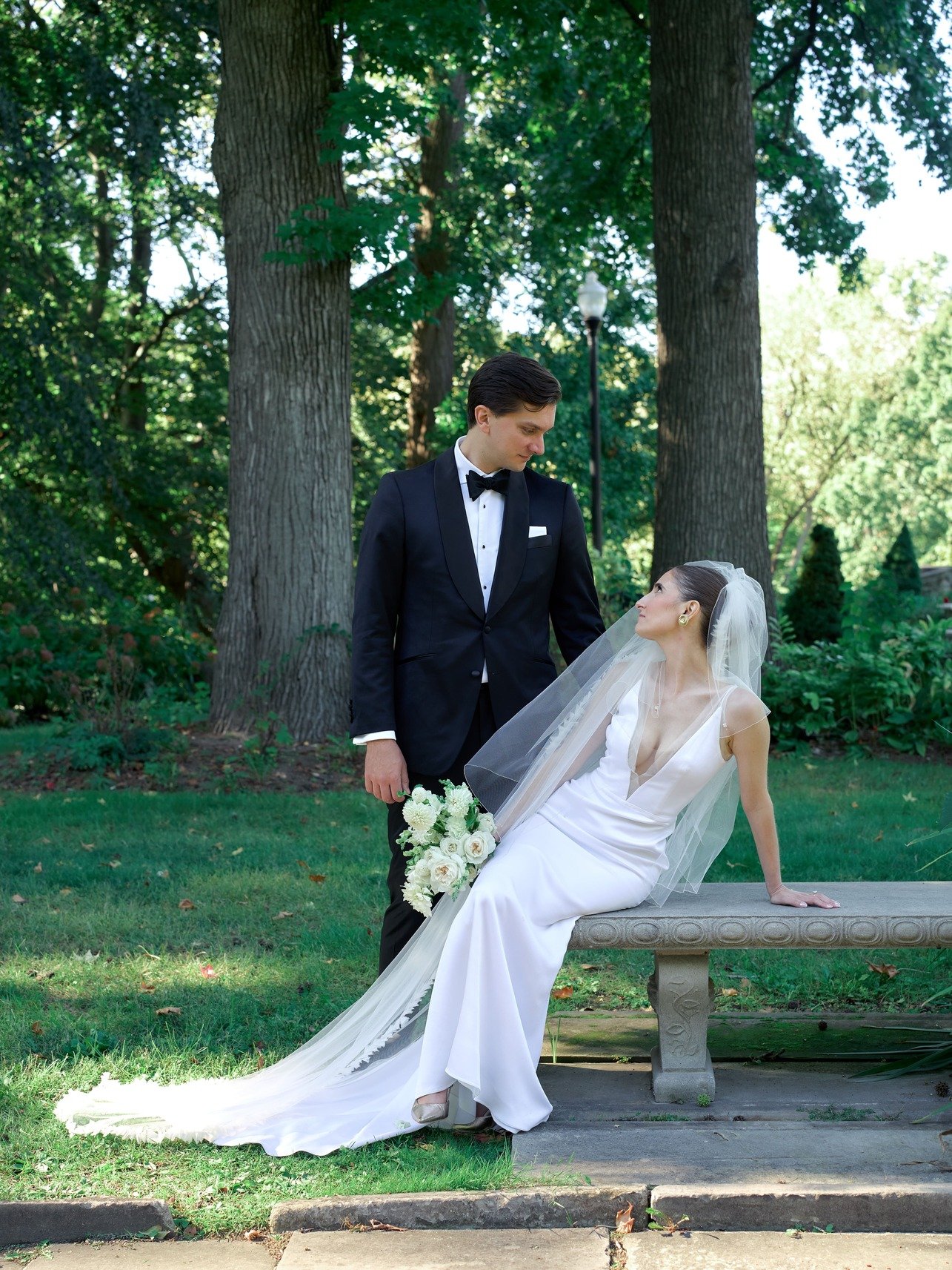 A bride and groom in wedding attire in a park, with large trees and greenery in the background. The bride is sitting on a stone bench holding a bouquet, and the groom is standing beside her, looking at her.