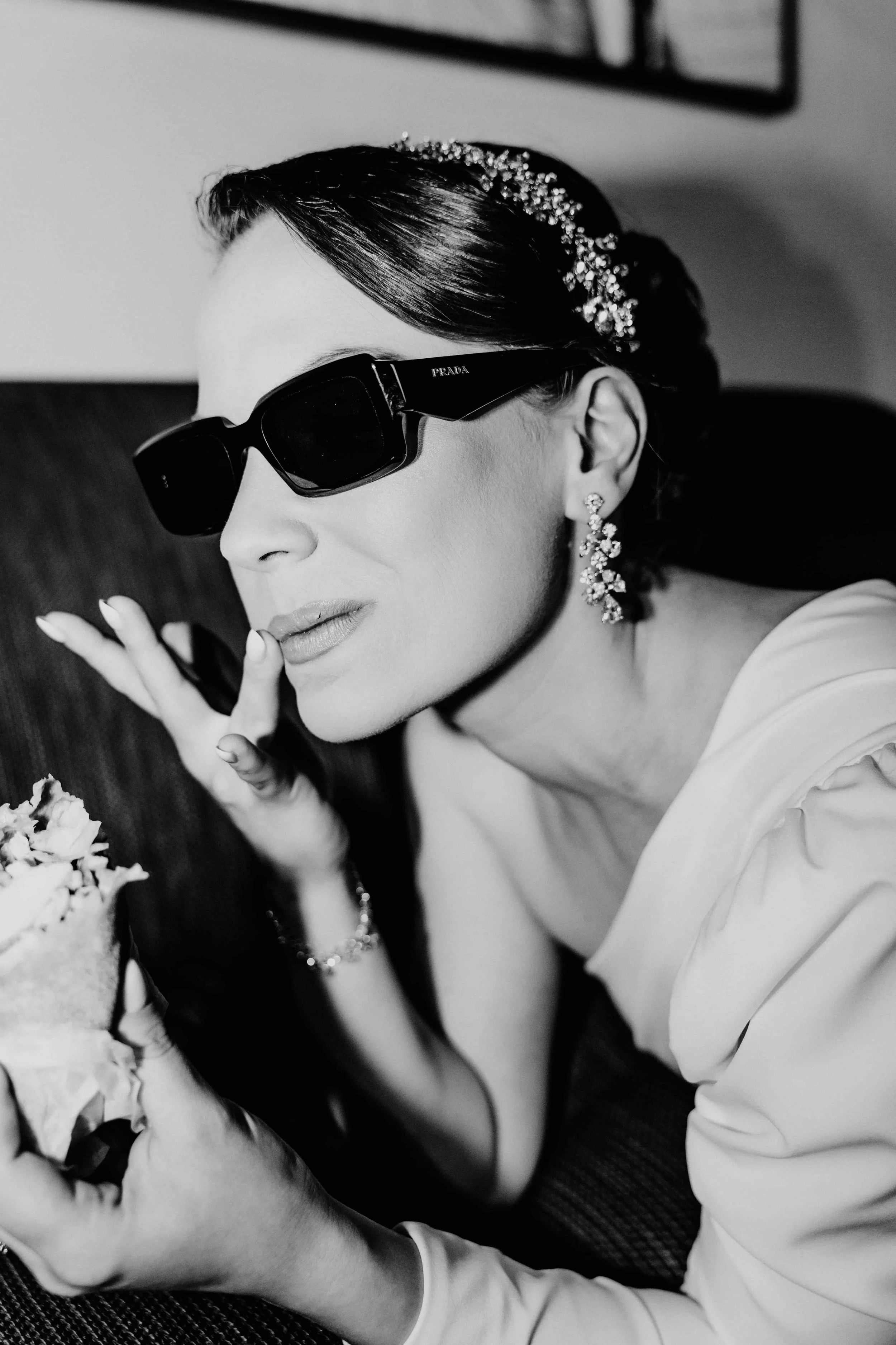 Black and white photo of a woman wearing large Prada sunglasses, elegant earrings, and a decorative headband, holding a cake and making a peace sign.