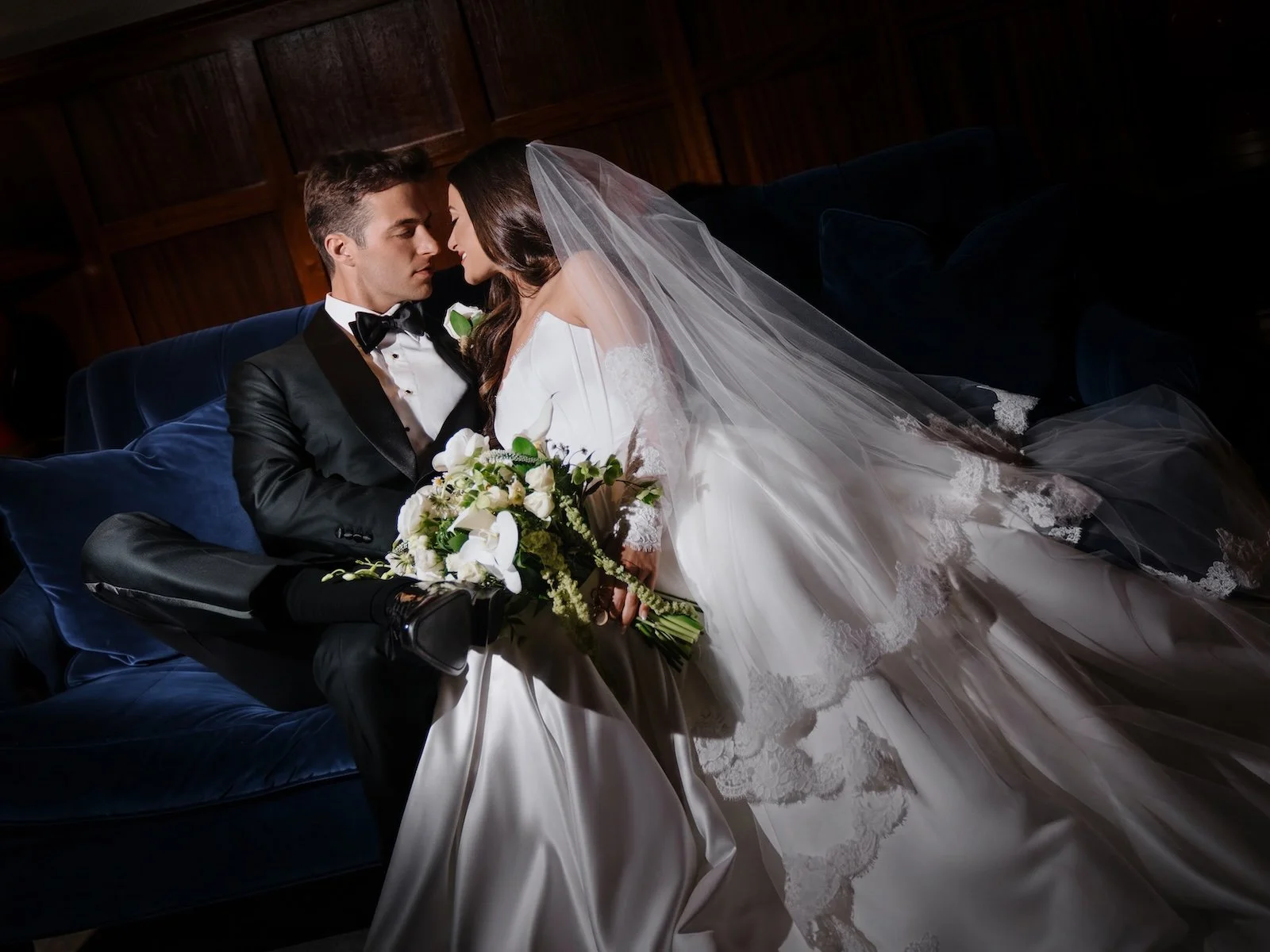 A bride and groom sitting closely together on a dark blue velvet couch, sharing an intimate moment. The groom is wearing a black tuxedo with a bowtie, and the bride is in a white wedding gown with a long veil. She is holding a bouquet of white flowers. The background features dark wooden wall paneling.