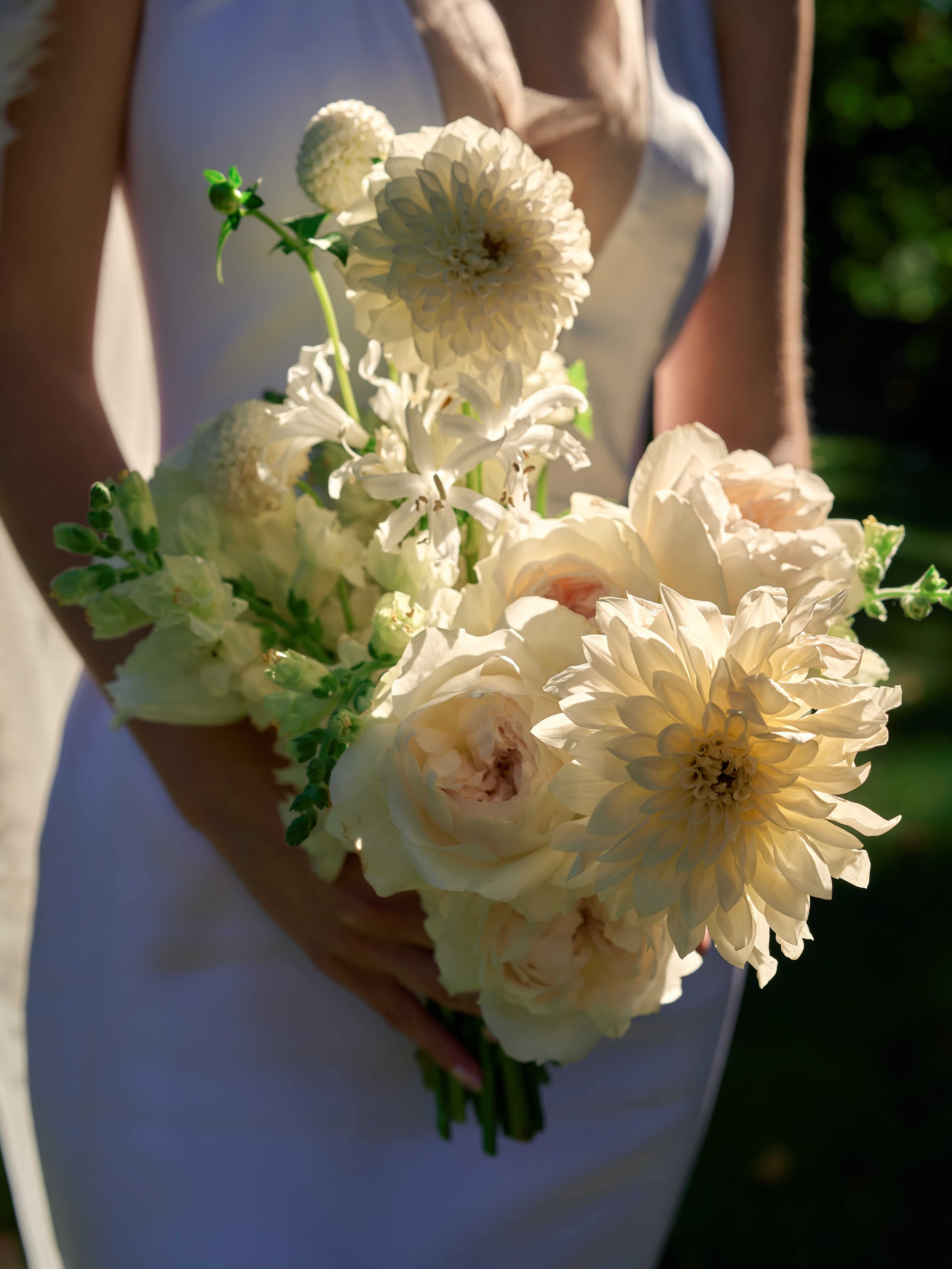 Woman in a white dress holding a bouquet of white flowers including dahlias and roses outside in sunlight.