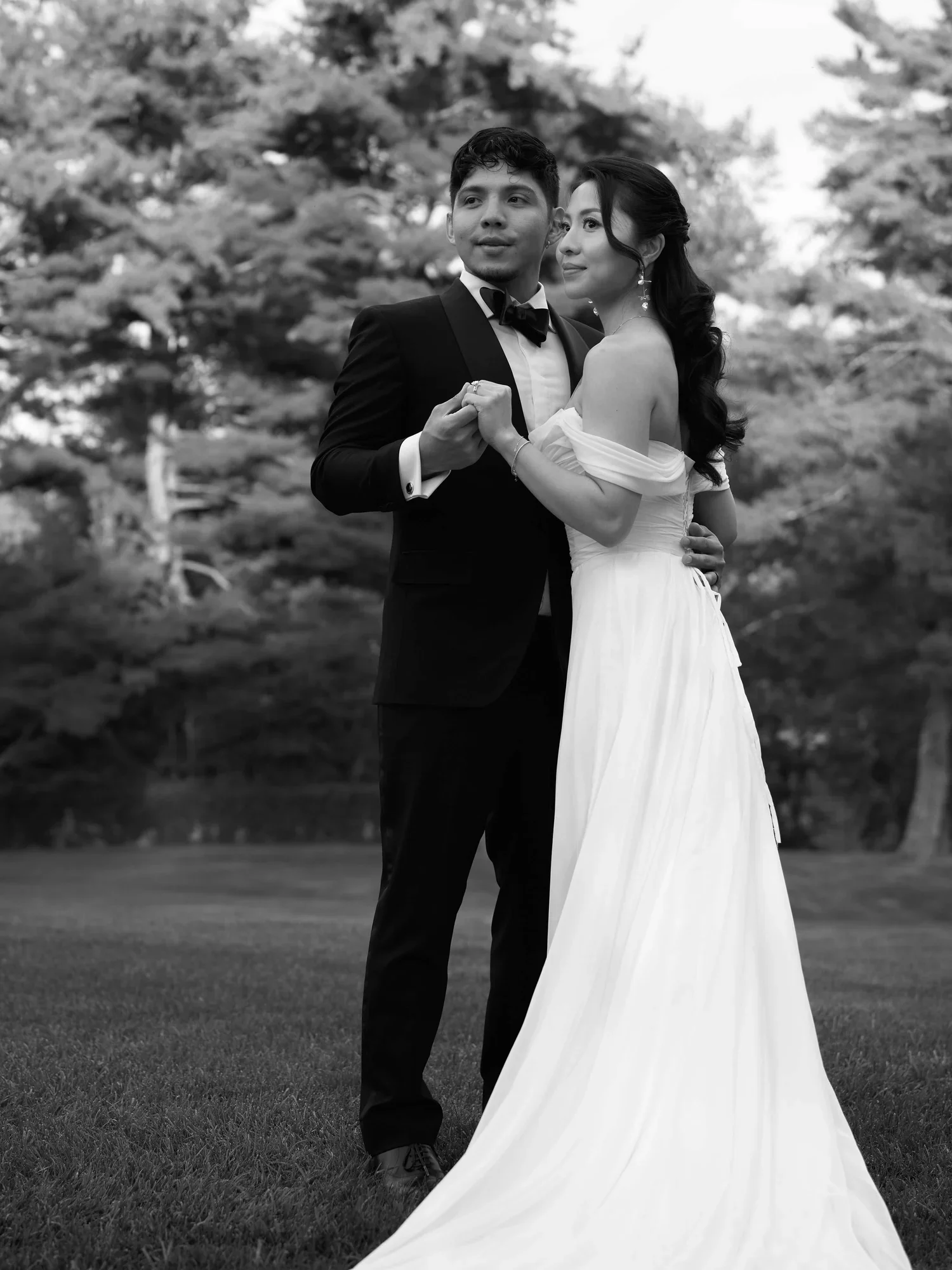 A black and white photo of a bride and groom dancing outdoors on grass with trees in the background. The groom wears a tuxedo, and the bride wears an off-shoulder wedding gown.