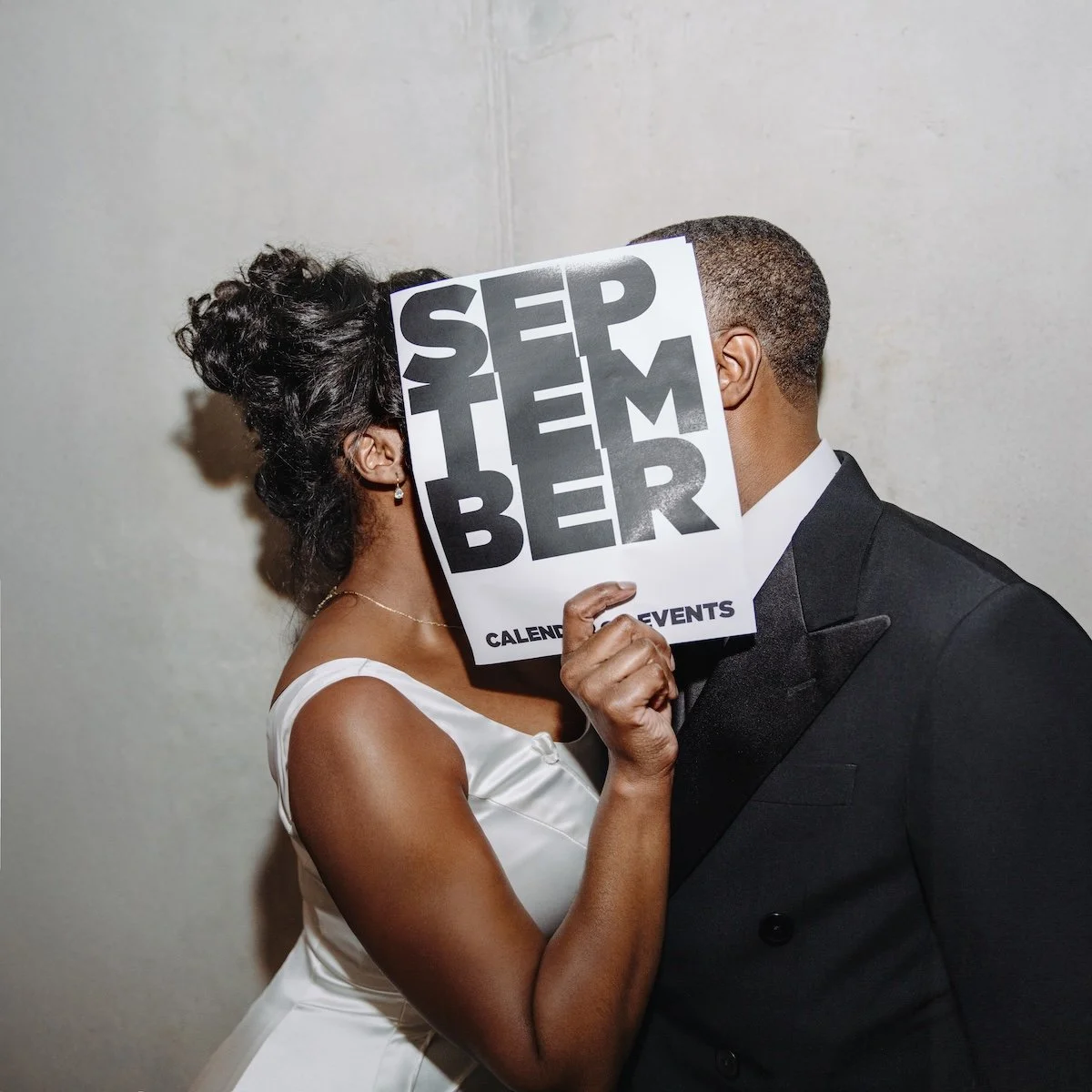 A woman in a white dress and a man in a black suit are facing each other with their faces close, holding a large sign with bold black and gray text that covers their faces. The sign reads 'SEPTEMBER' and 'CALENDAR OF EVENTS'.