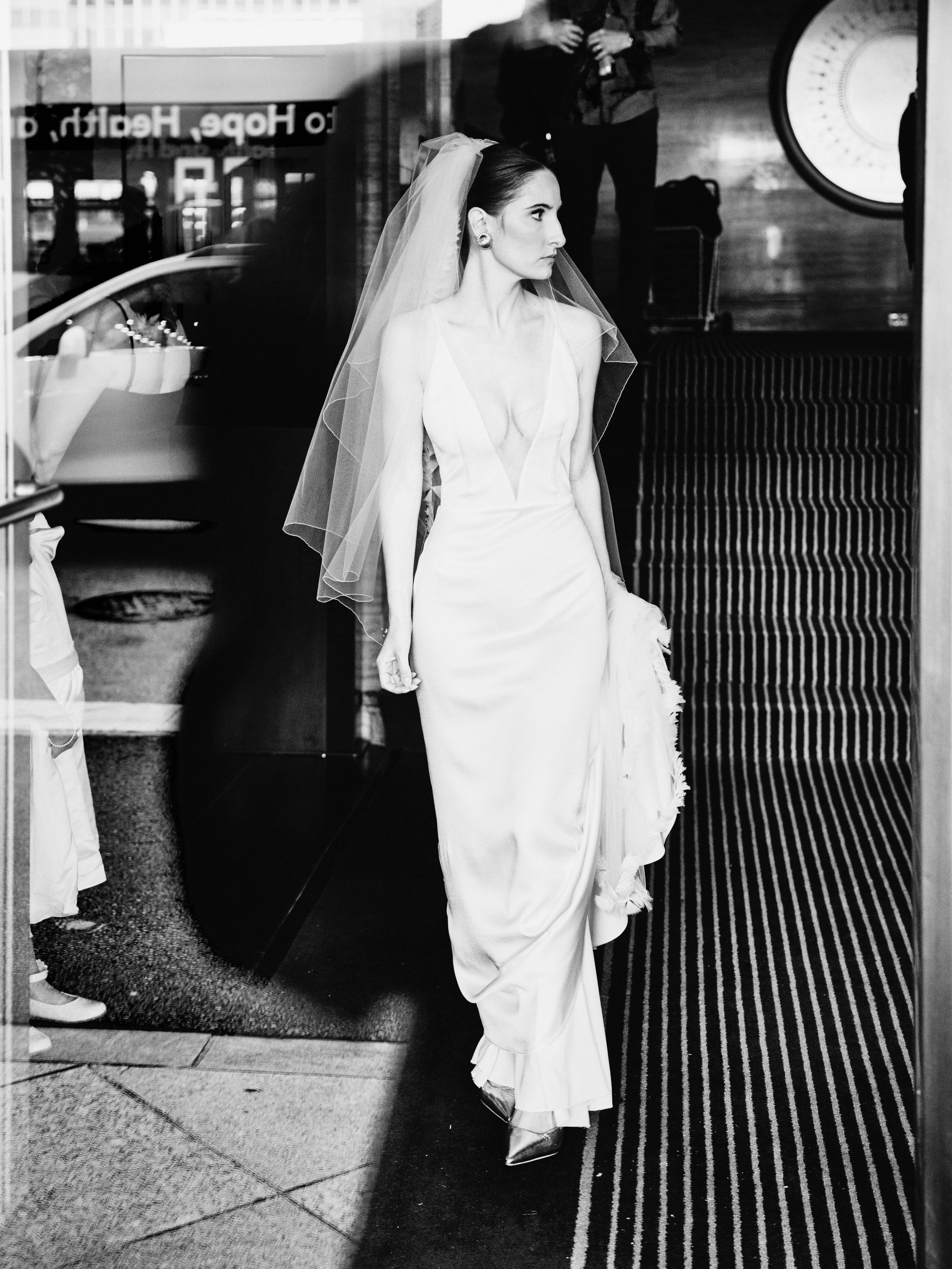 Black and white photo of a bride in a wedding dress and veil walking through a glass door, with her reflection visible on the glass.