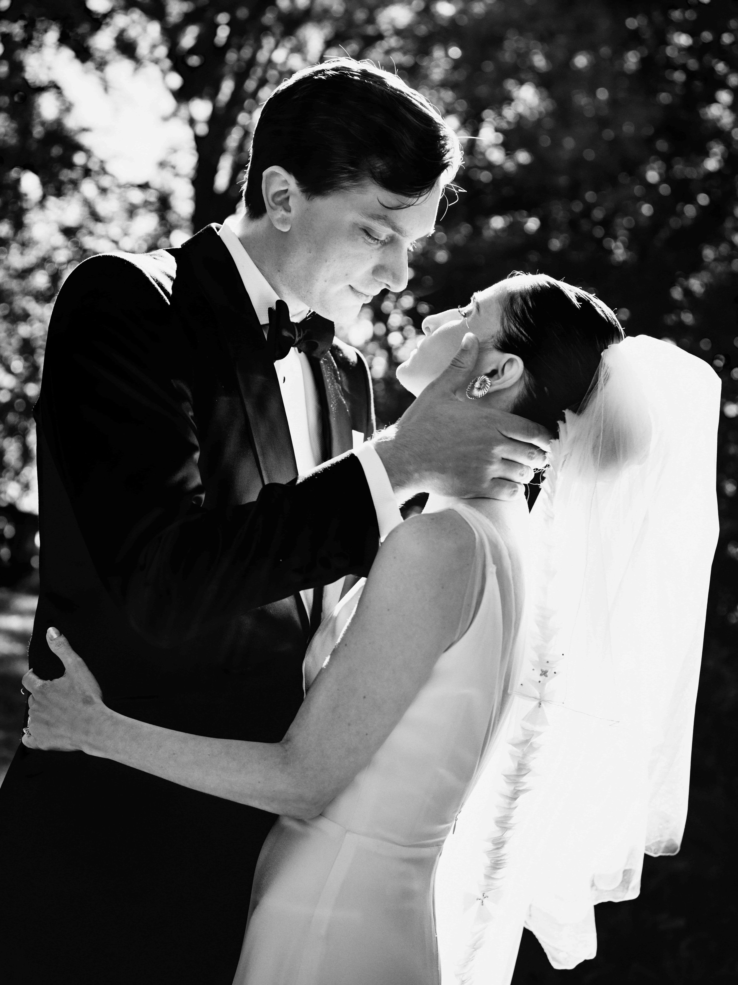 Black and white photo of a groom gently holding and looking at a bride's face, with sunlight filtering through trees in the background.
