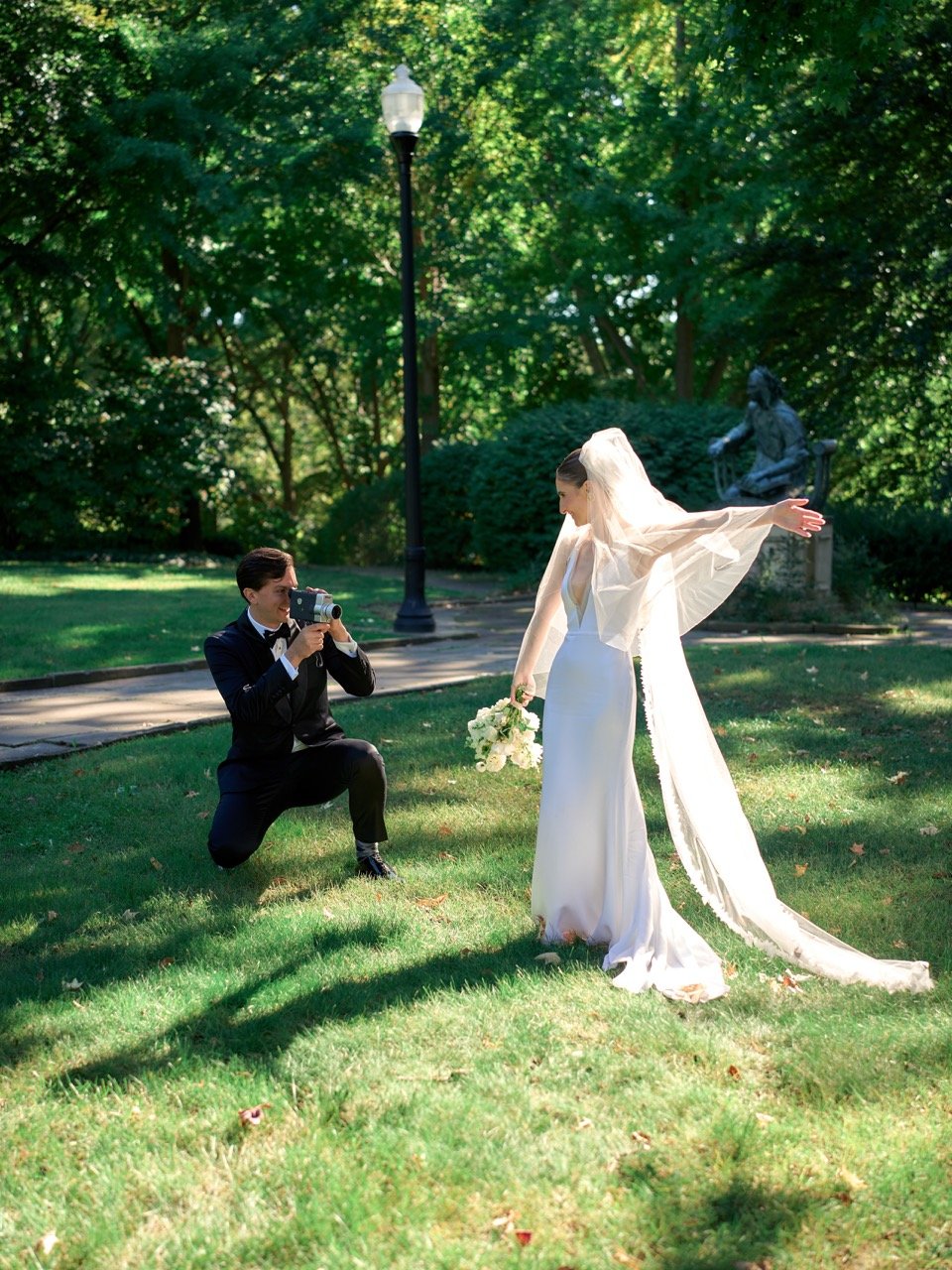 A groom in a black tuxedo kneeling on one knee, taking a photo of a bride in a white wedding dress with a veil and holding a bouquet, in a lush green park with a statue and a lamppost in the background.