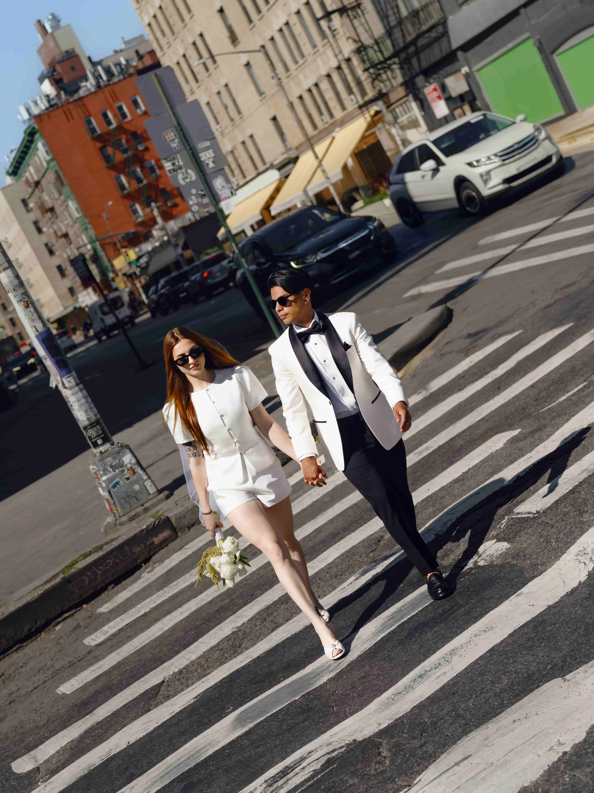 A stylish couple dressed in formal wedding attire walking hand in hand across a city street crosswalk on a sunny day.