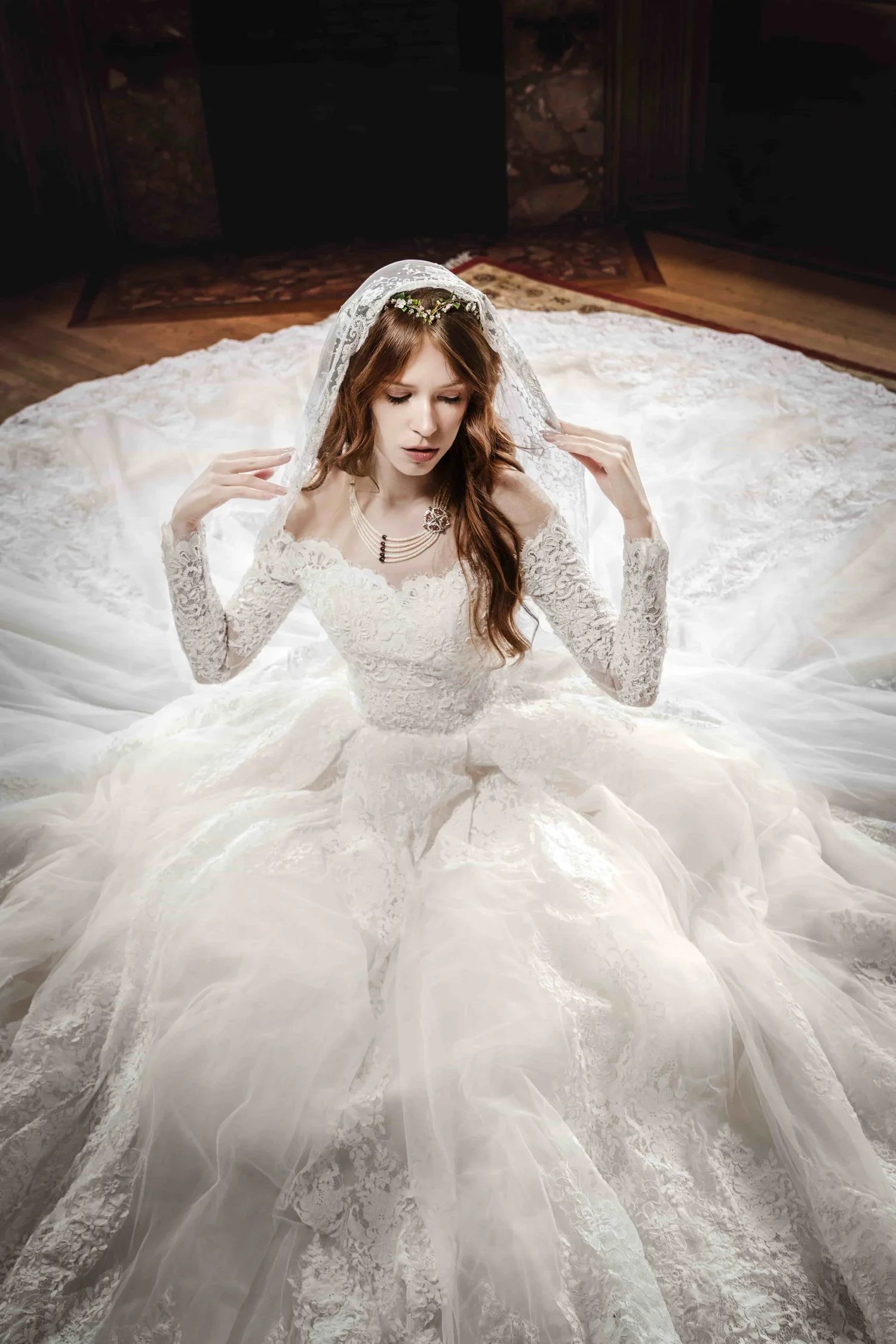 A bride in a white lace wedding dress with long sleeves, sitting on a wooden floor, holding her veil, with a dark background and a patterned carpet behind her.