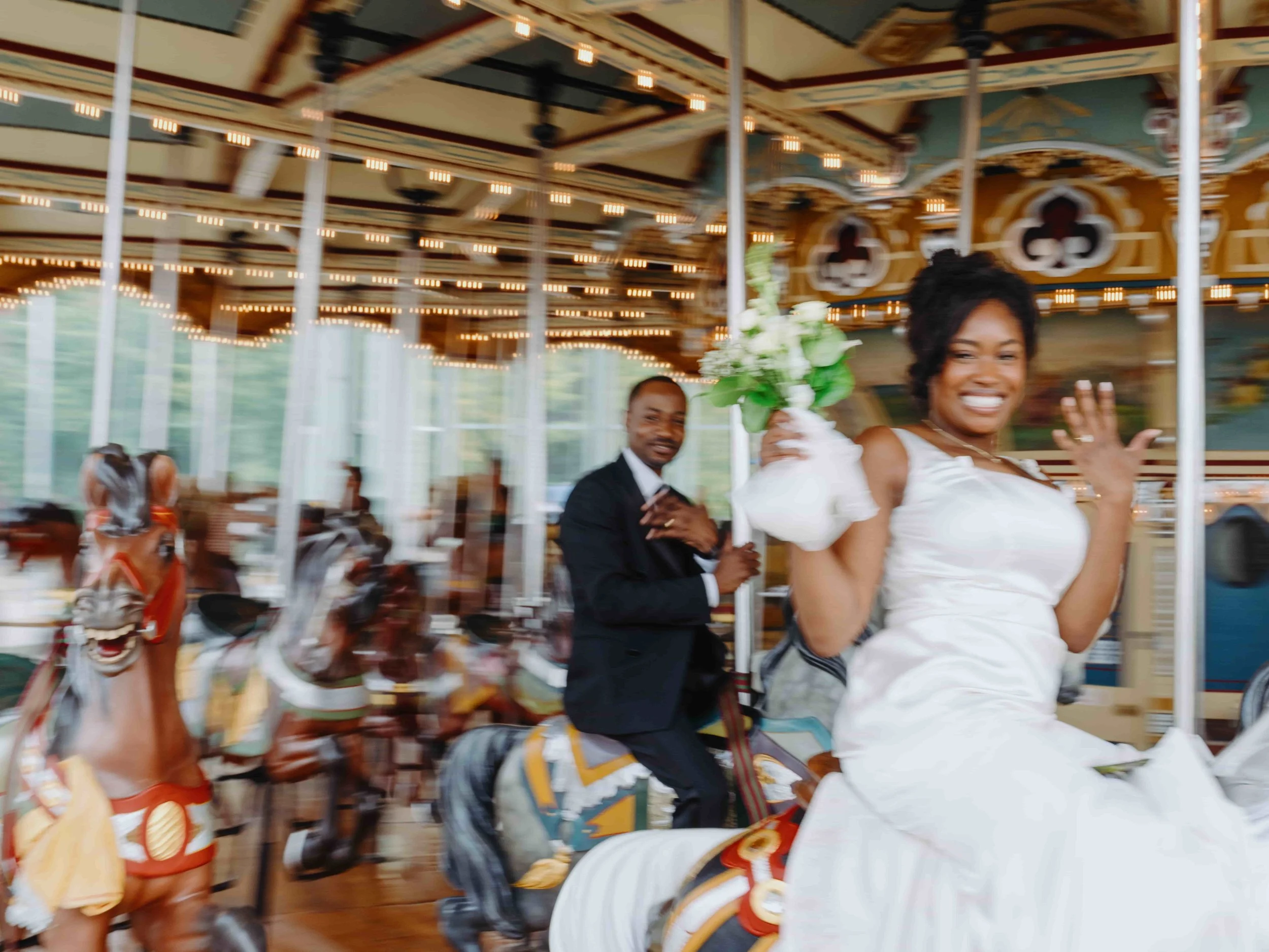 People riding a carousel with carousel horses, one woman smiling and holding a bouquet, a man in a suit also riding, all indoors with hanging lights.
