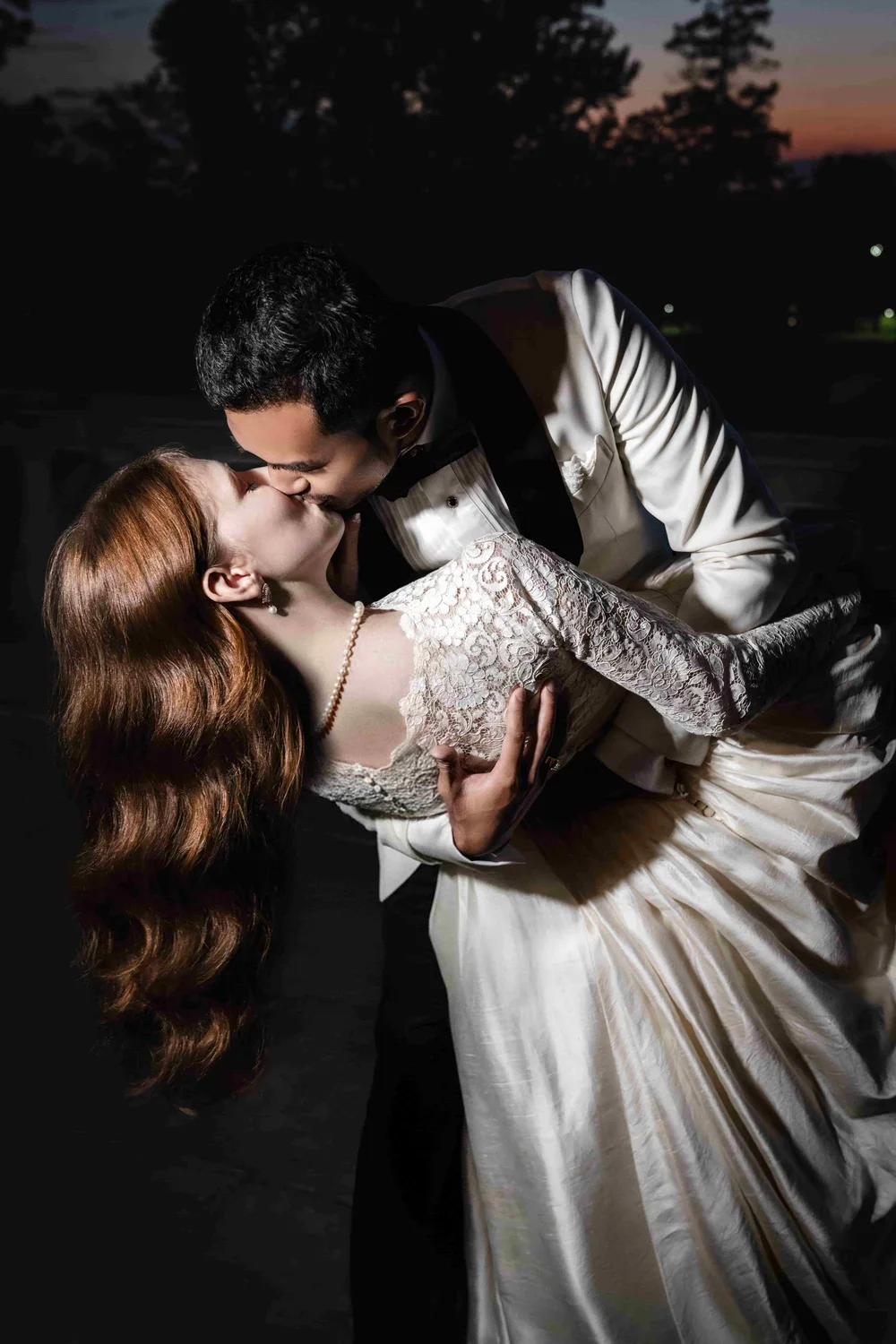 A couple in formal wedding attire sharing a kiss outdoors at sunset.