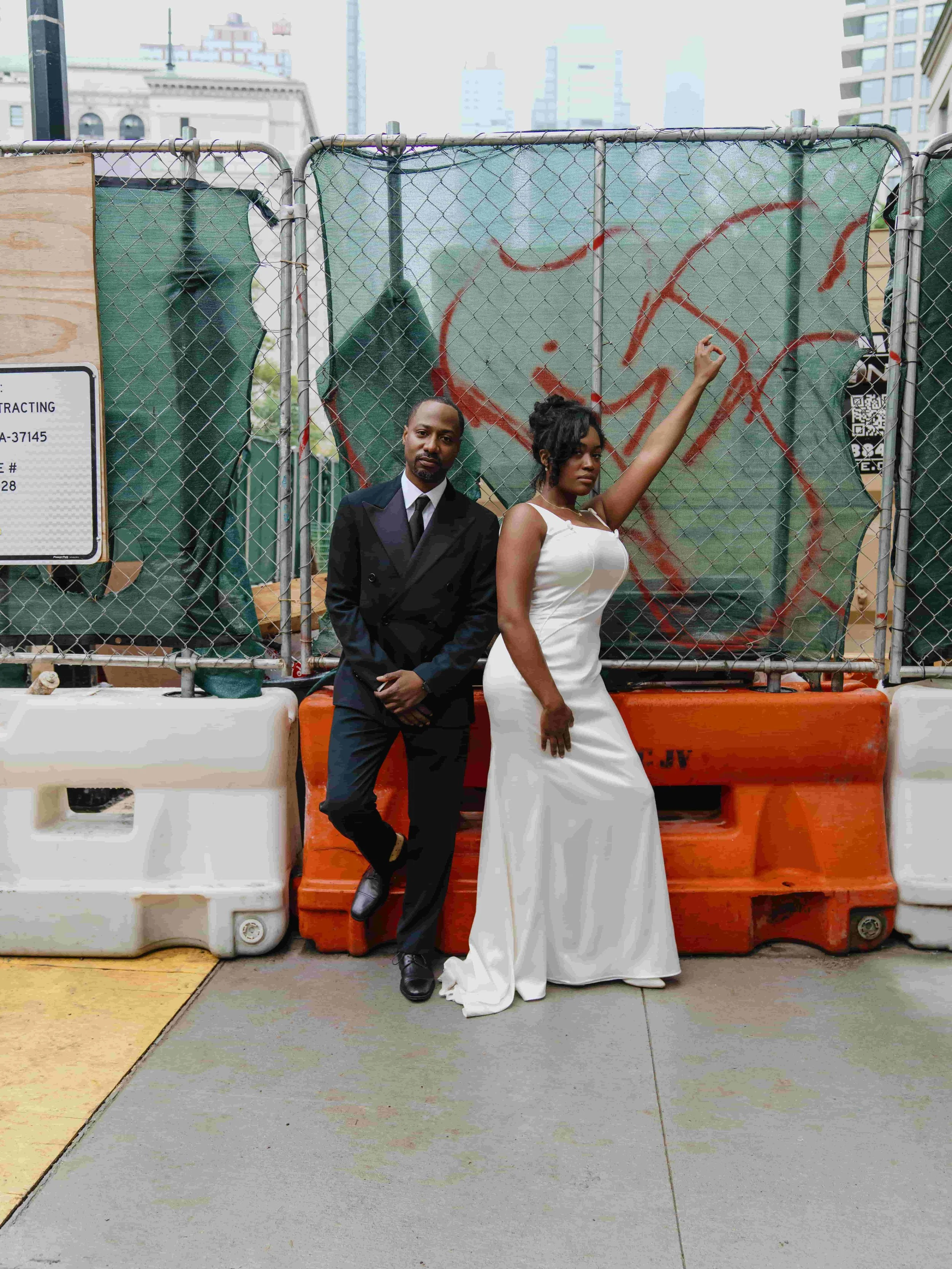 A man in a black suit and a woman in a white dress standing in front of a construction barrier on an urban street, with city buildings in the background.