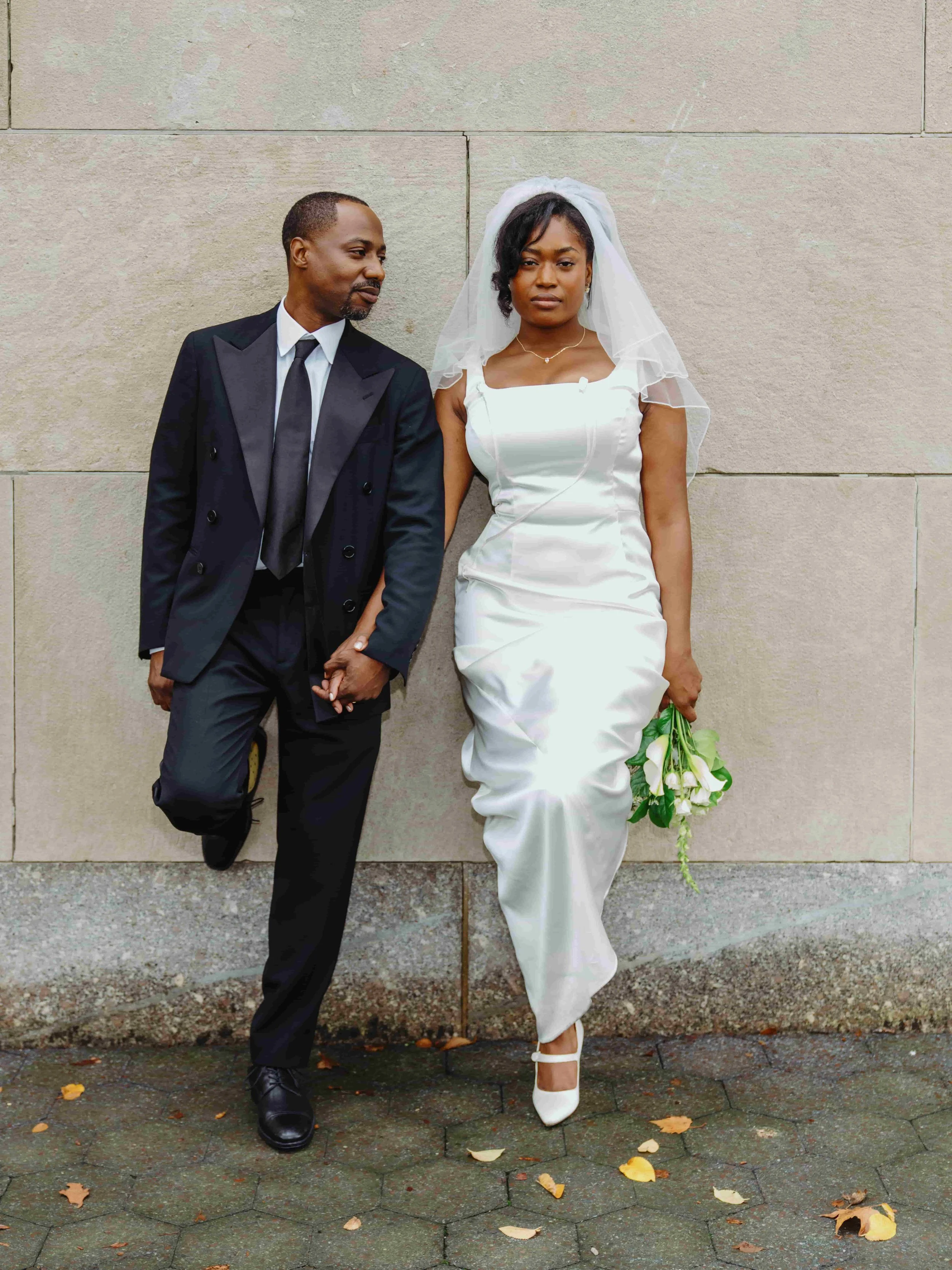 A couple dressed in wedding attire standing against a stone wall. The woman is in a white wedding dress with a veil, holding a bouquet of white flowers. The man is in a black suit with a tie, standing beside her.