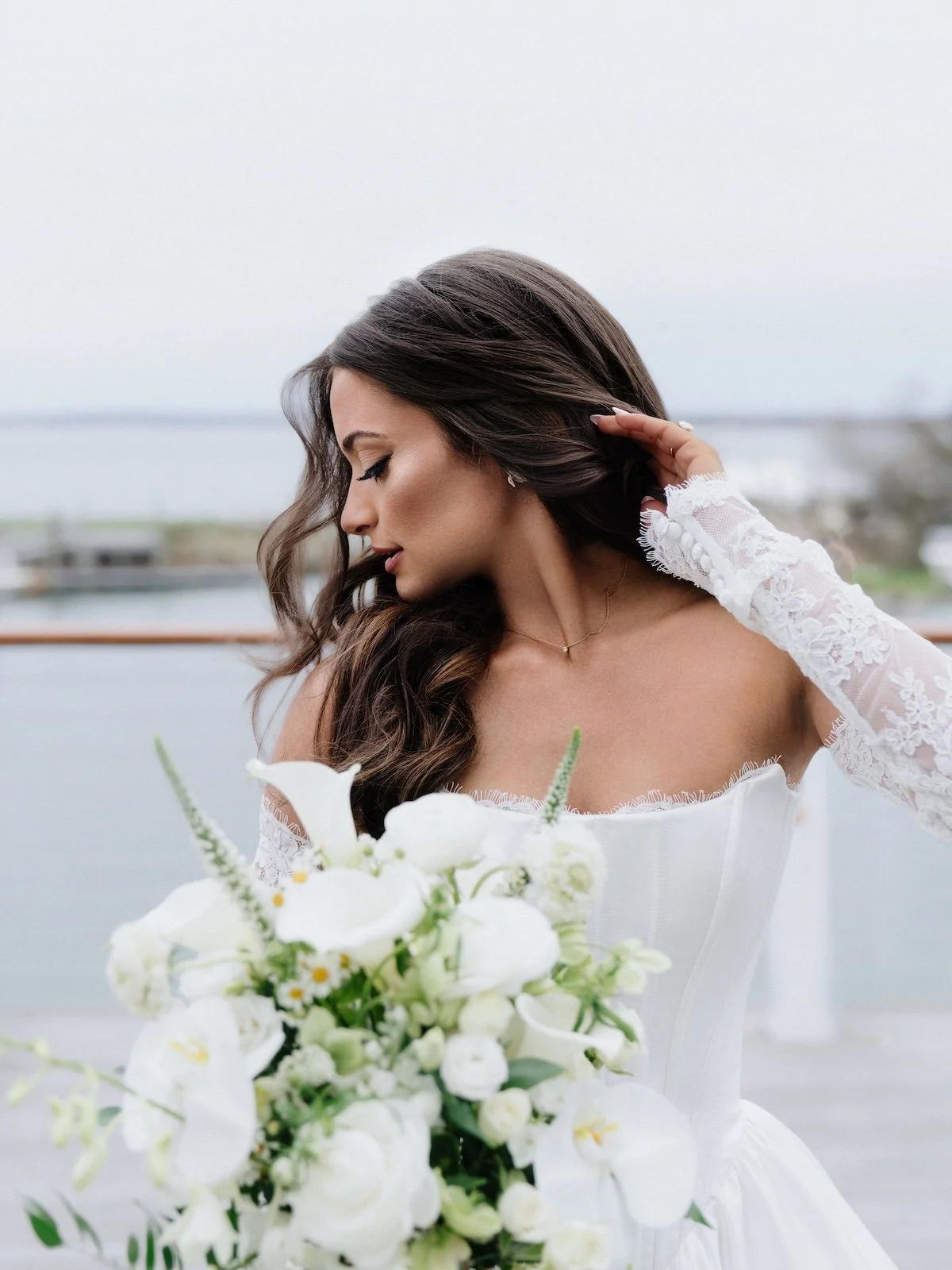 A bride in a white wedding dress holding a bouquet of white flowers, with long brown hair, standing outdoors near a body of water under a cloudy sky.