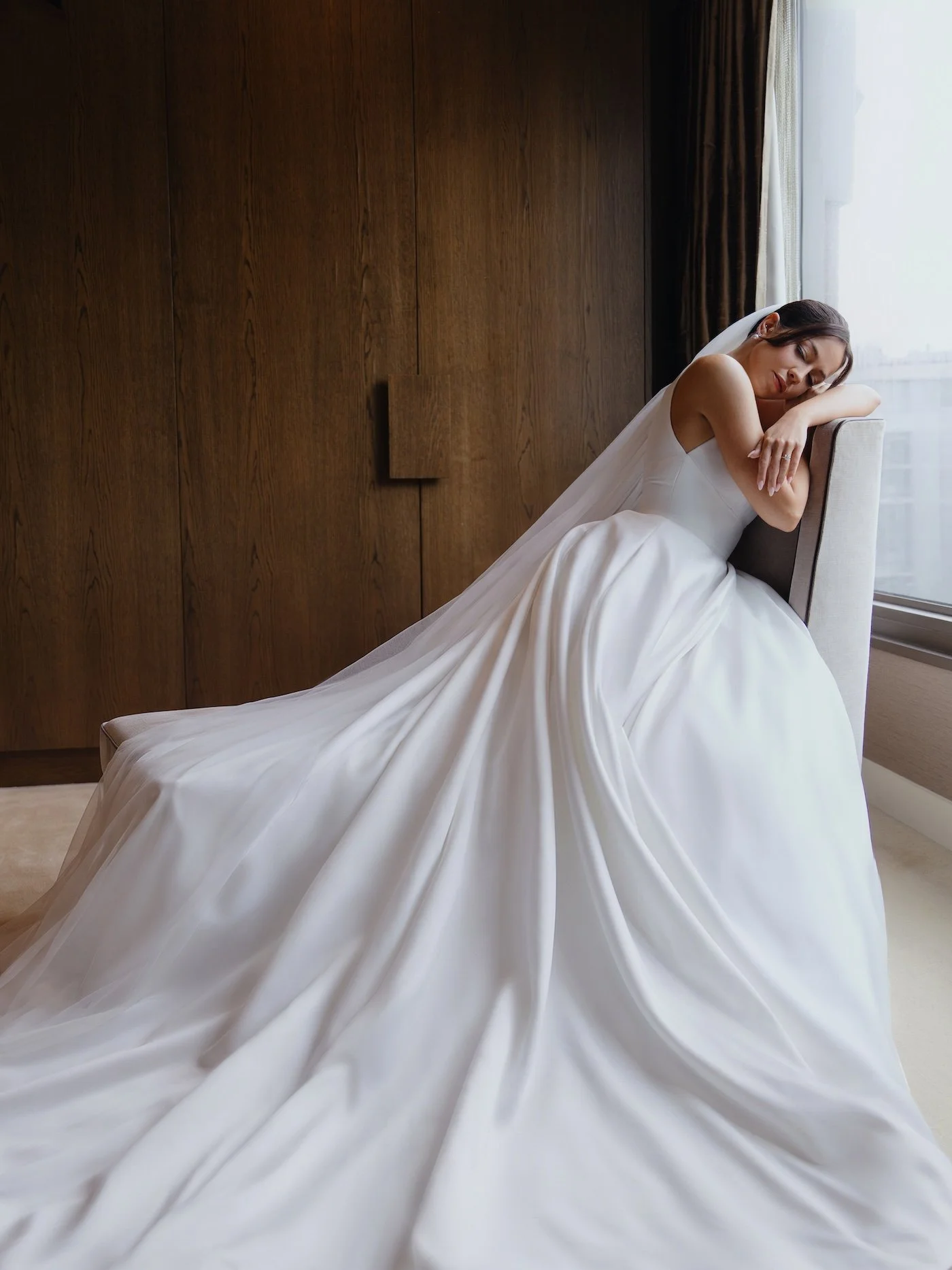 A bride in a white wedding dress is sitting on a chair, resting her head on her hands, by a window with natural light. She looks peaceful and relaxed.